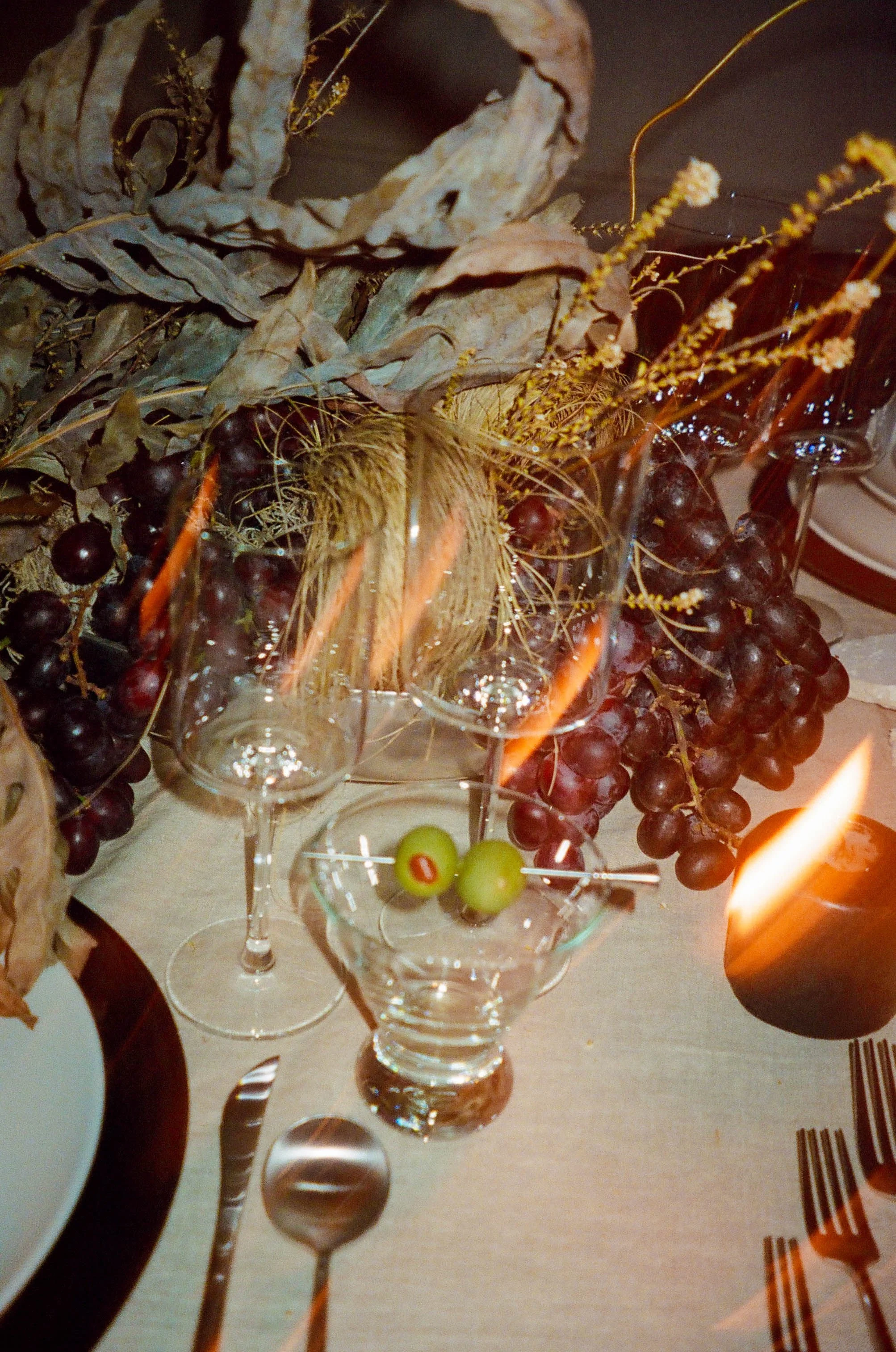 A table with dried leaves, grapes, a candle, wine glasses, and a centerpiece with dried flowers and fruits.