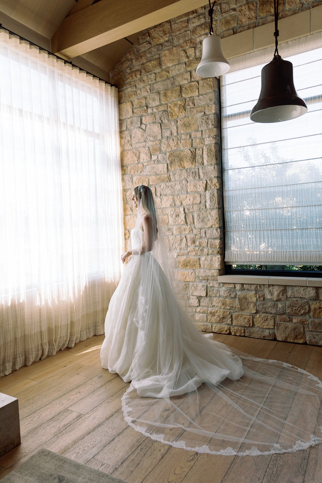 A bride in a wedding dress standing by a window with sheer curtains, in a room with a stone wall and wooden floors, illuminated by natural light.