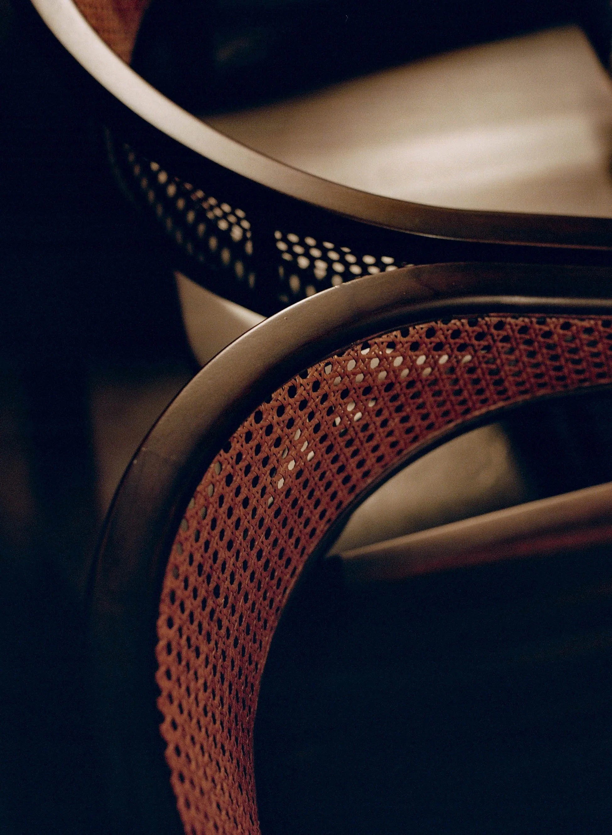Close-up of two chairs with woven cane backs, one with a reddish hue and the other with a dark finish, in a warm lighting setting.