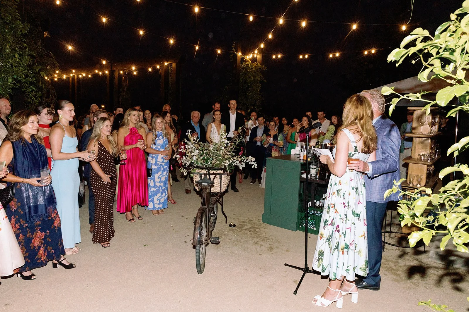 A wedding reception outdoors at night with string lights overhead. The bride and groom are speaking into a microphone while guests, dressed in formal attire, listen. There is a green bar and a bicycle with flowers in the foreground.
