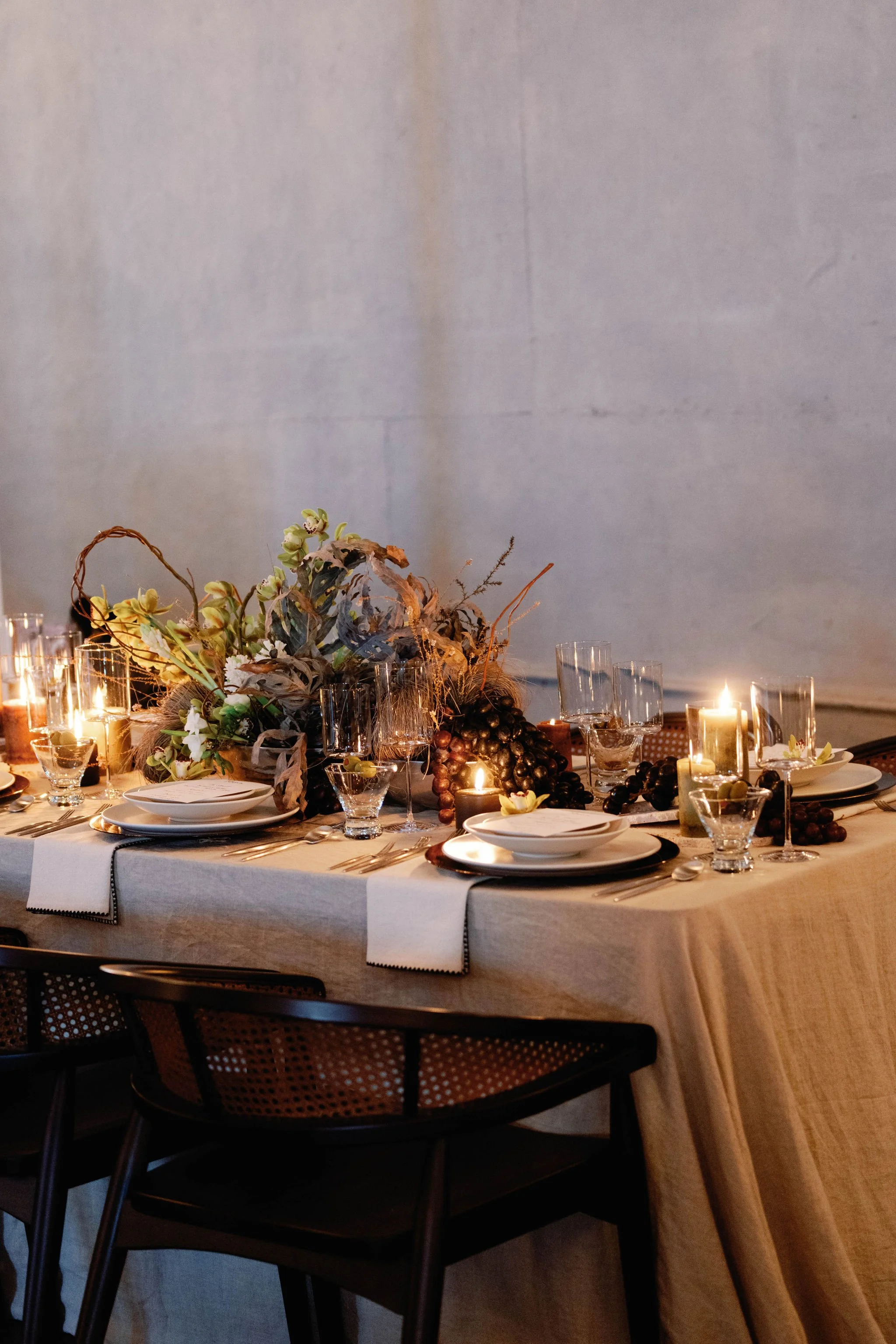 Elegant dining table with candles, floral arrangement, glassware, and plates, set for a special occasion, against a plain wall background.