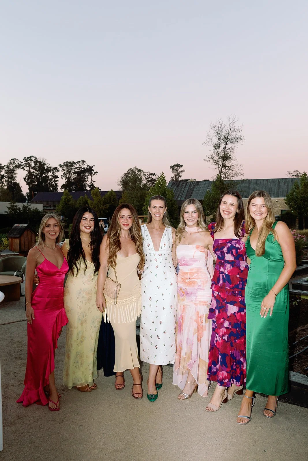 Seven women in colorful dresses standing together outdoors at sunset, smiling at the camera.