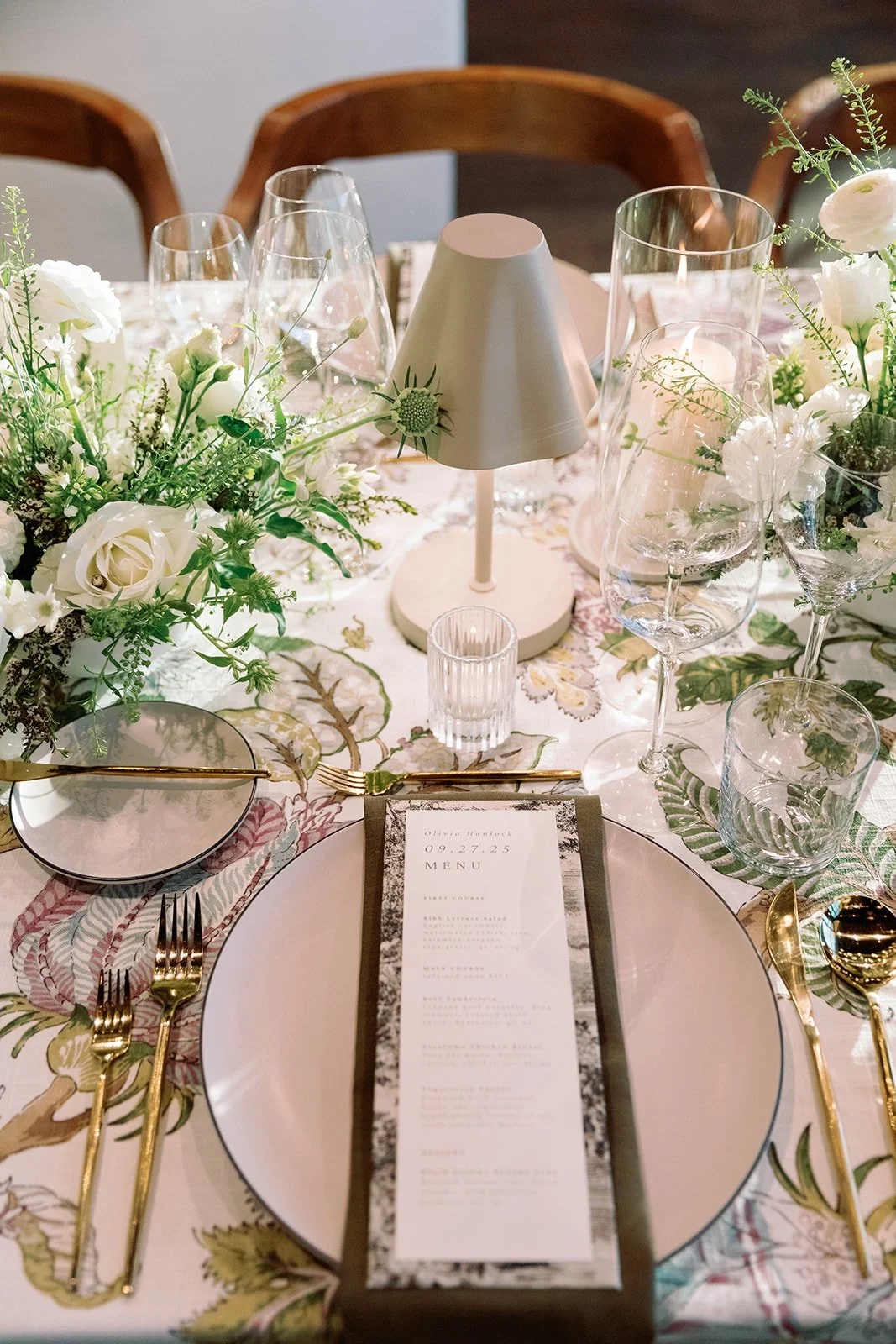 A beautifully decorated table set for a wedding or formal event with white flowers, gold cutlery, glassware, a menu, and a small lamp in the center.
