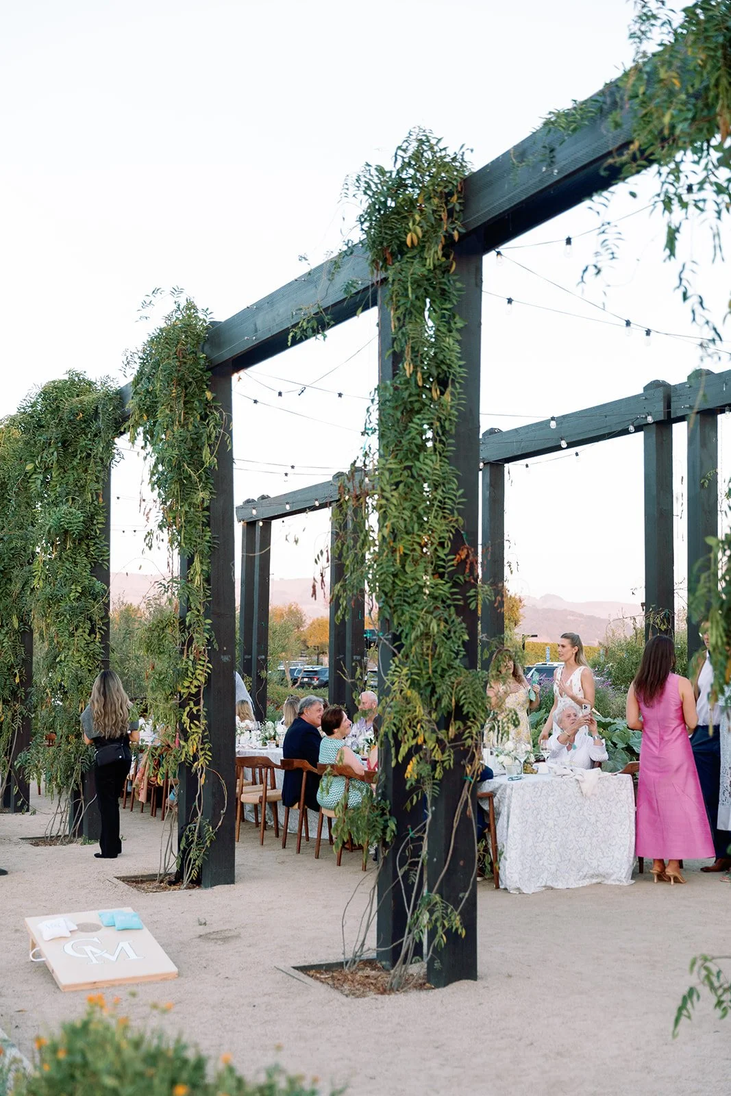 Outdoor wedding reception with guests sitting at tables under a wooden pergola decorated with greenery and string lights.