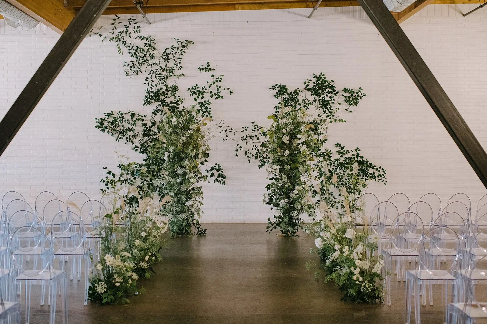 An indoor wedding aisle with a white brick wall background, decorated with large floral arrangements of green leaves and white flowers along the sides, leading to a floral arch.