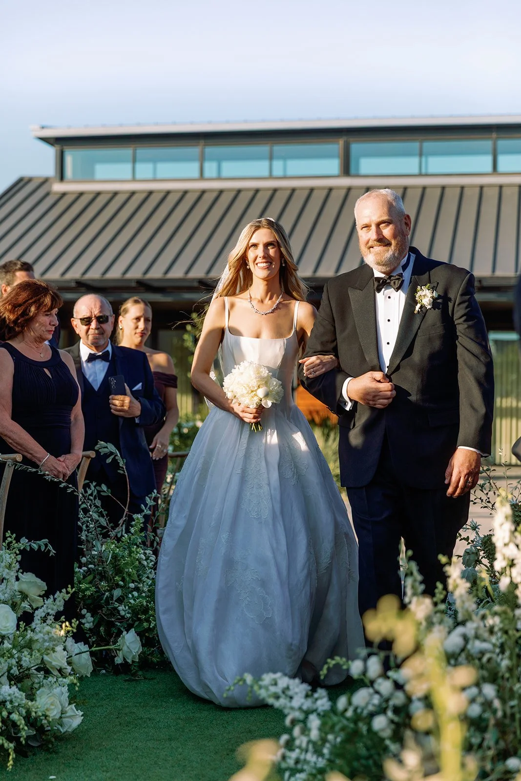 A bride walking down the aisle with her father during a wedding ceremony outdoors, surrounded by guests and floral decorations.