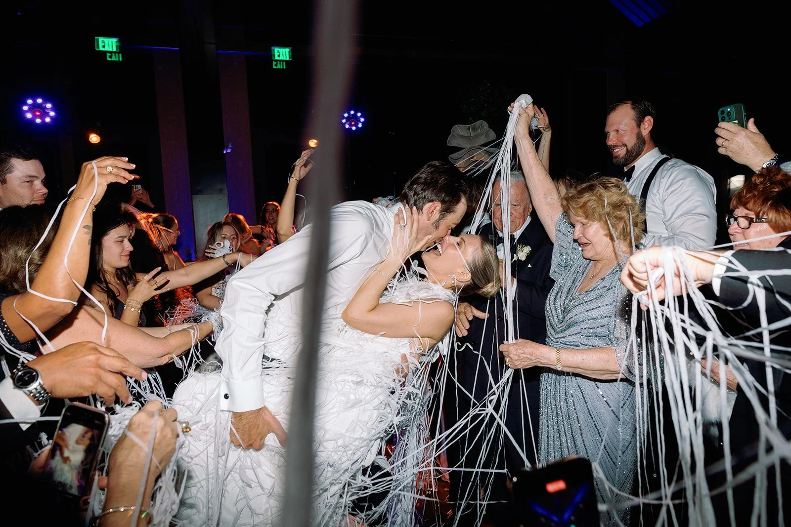 A bride and groom share a kiss while surrounded by family and friends at a wedding reception, with streamers in the air celebrating their union.