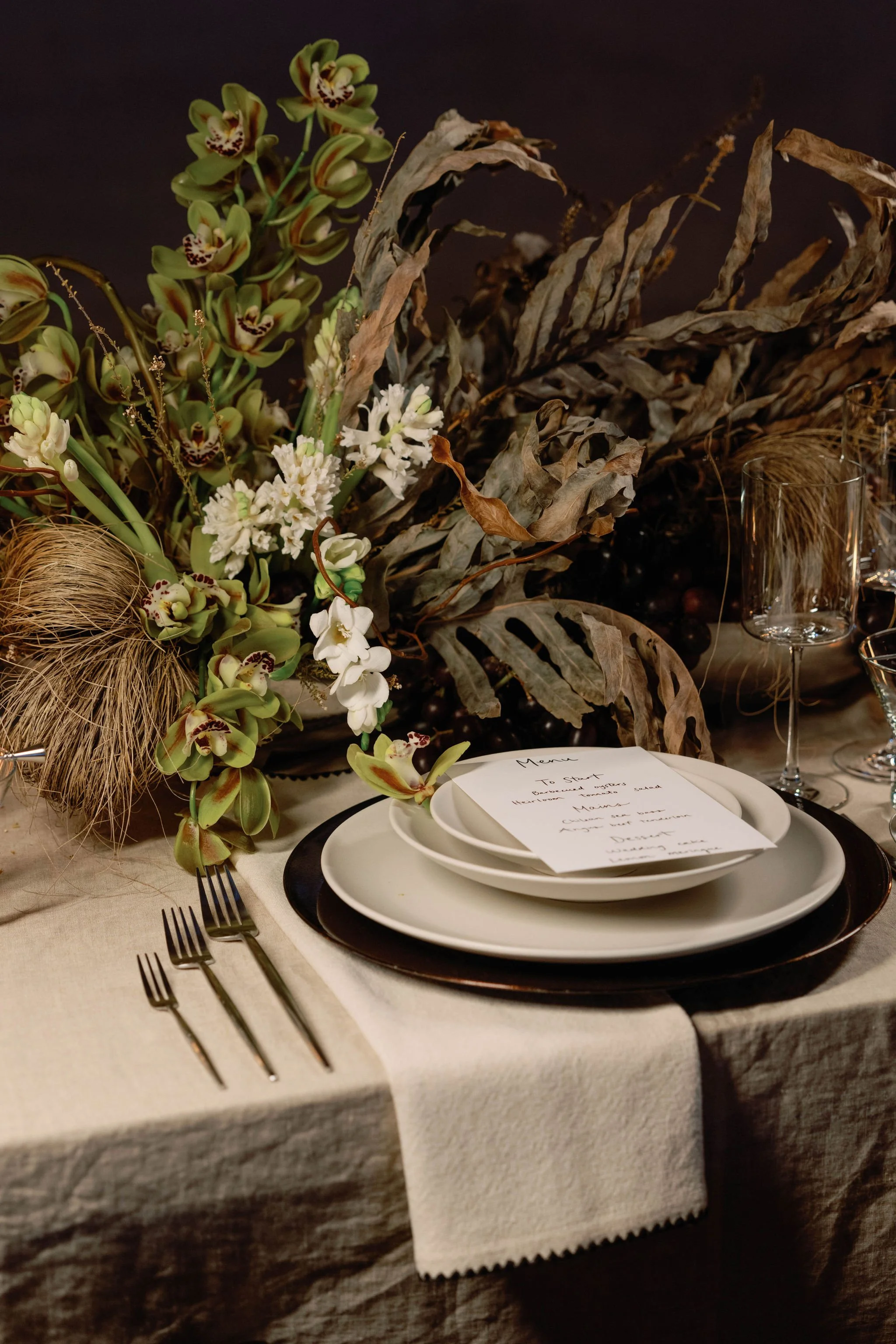 Elegant table setting with a decorative floral centerpiece consisting of orchids and dried leaves, white and black plates, a handwritten menu card, three forks, and empty wine glasses on a beige tablecloth.