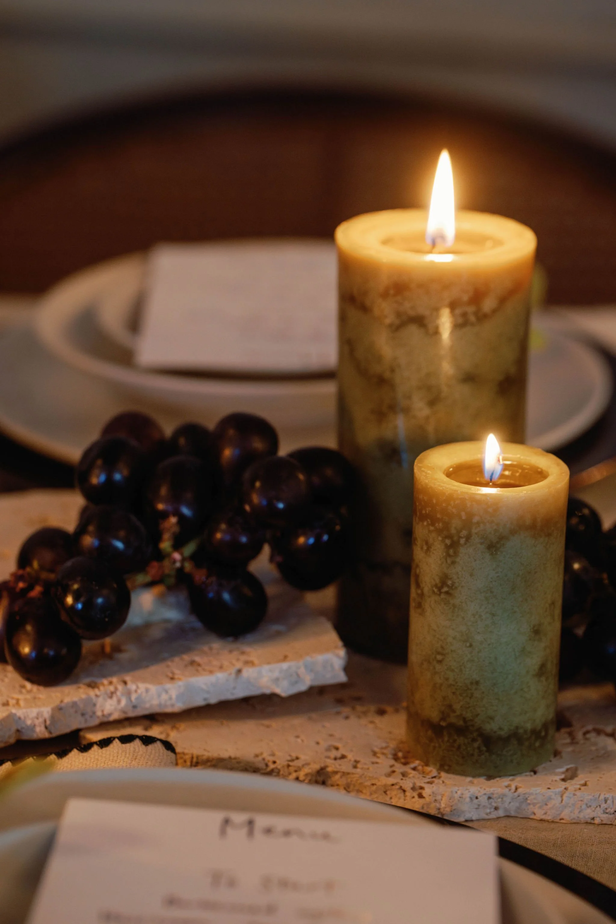 Decorative table setting featuring two lit pillar candles, a bunch of dark grapes, and a plate with a napkin and a card, in a warm, cozy ambiance.