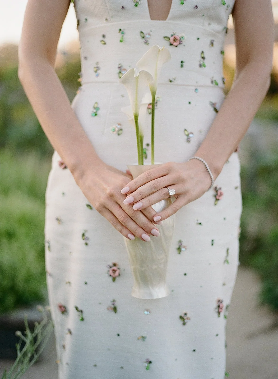 A woman in a white dress with floral embroidery holds a vase with three white calla lilies.
