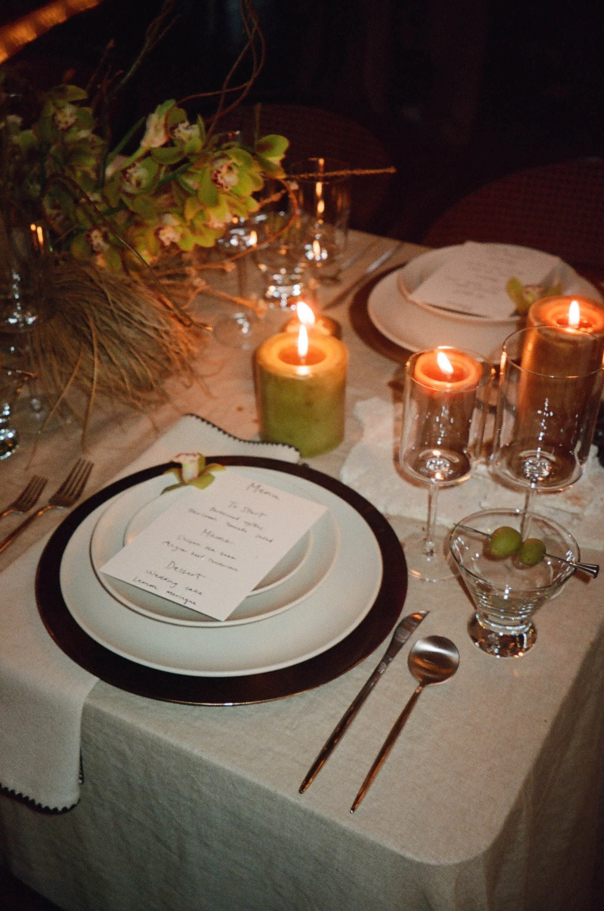 An elegant dining table set for a meal with white plates, a menu card, silverware, and a small bowl with green olives. The table is decorated with candles in green and clear glass holders, and there's a floral arrangement with green and beige flowers