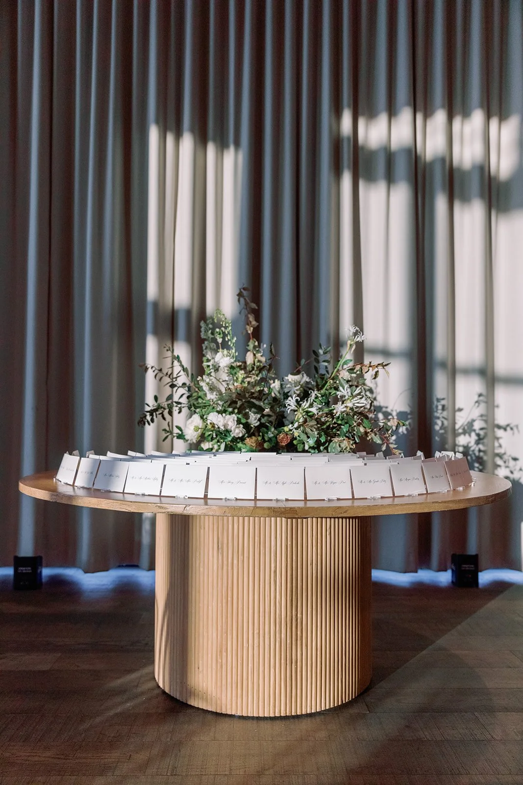 A round wooden table with a floral arrangement in the center, surrounded by small white cards, situated in front of a window with blinds.