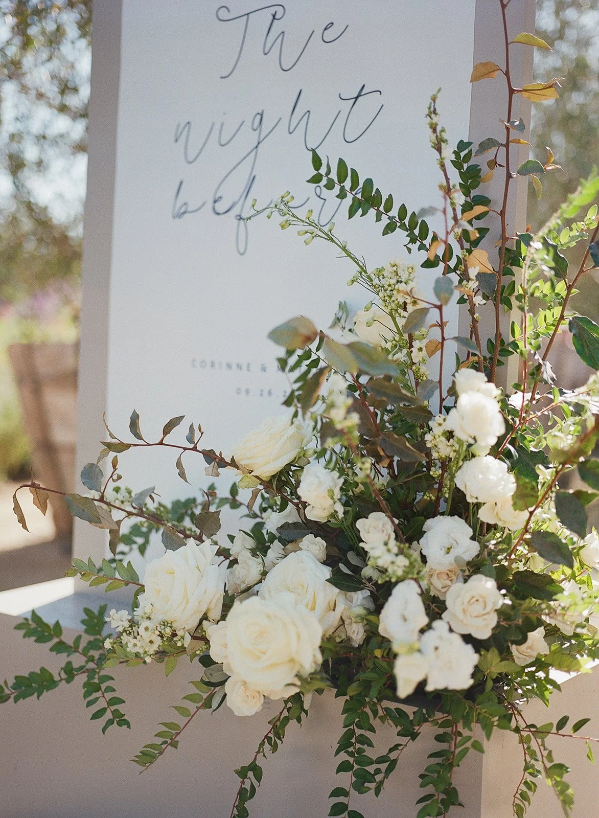 A white floral arrangement with roses and greenery in front of a white sign with cursive writing.