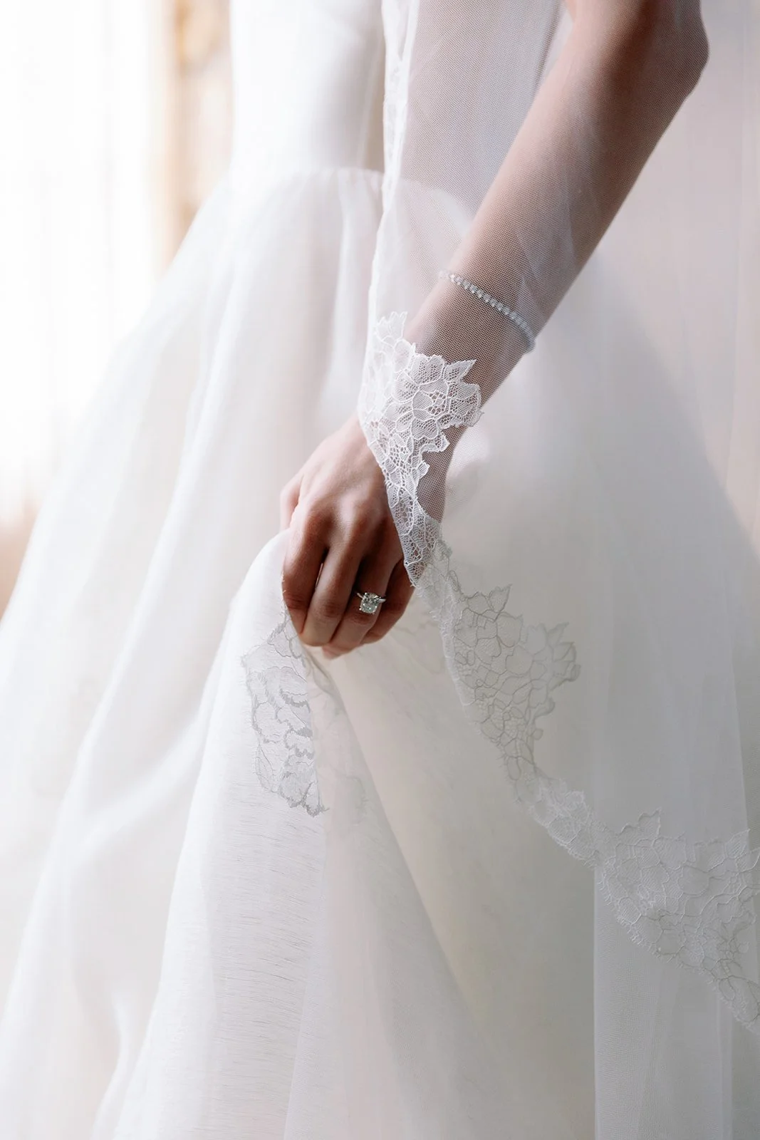 Close-up of a bride in a white wedding dress with lace sleeve details, her hand resting on her dress, wearing an engagement ring.