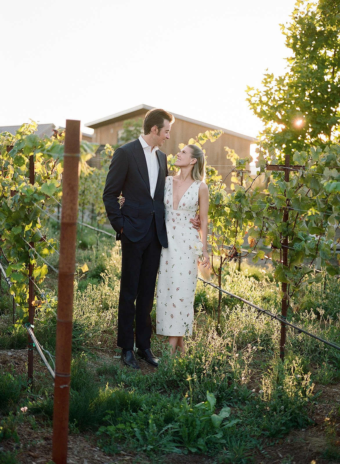 A couple standing close together in a vineyard during sunset, looking at each other lovingly. The man is dressed in a black suit, and the woman is wearing a floral white dress, holding a small bouquet.