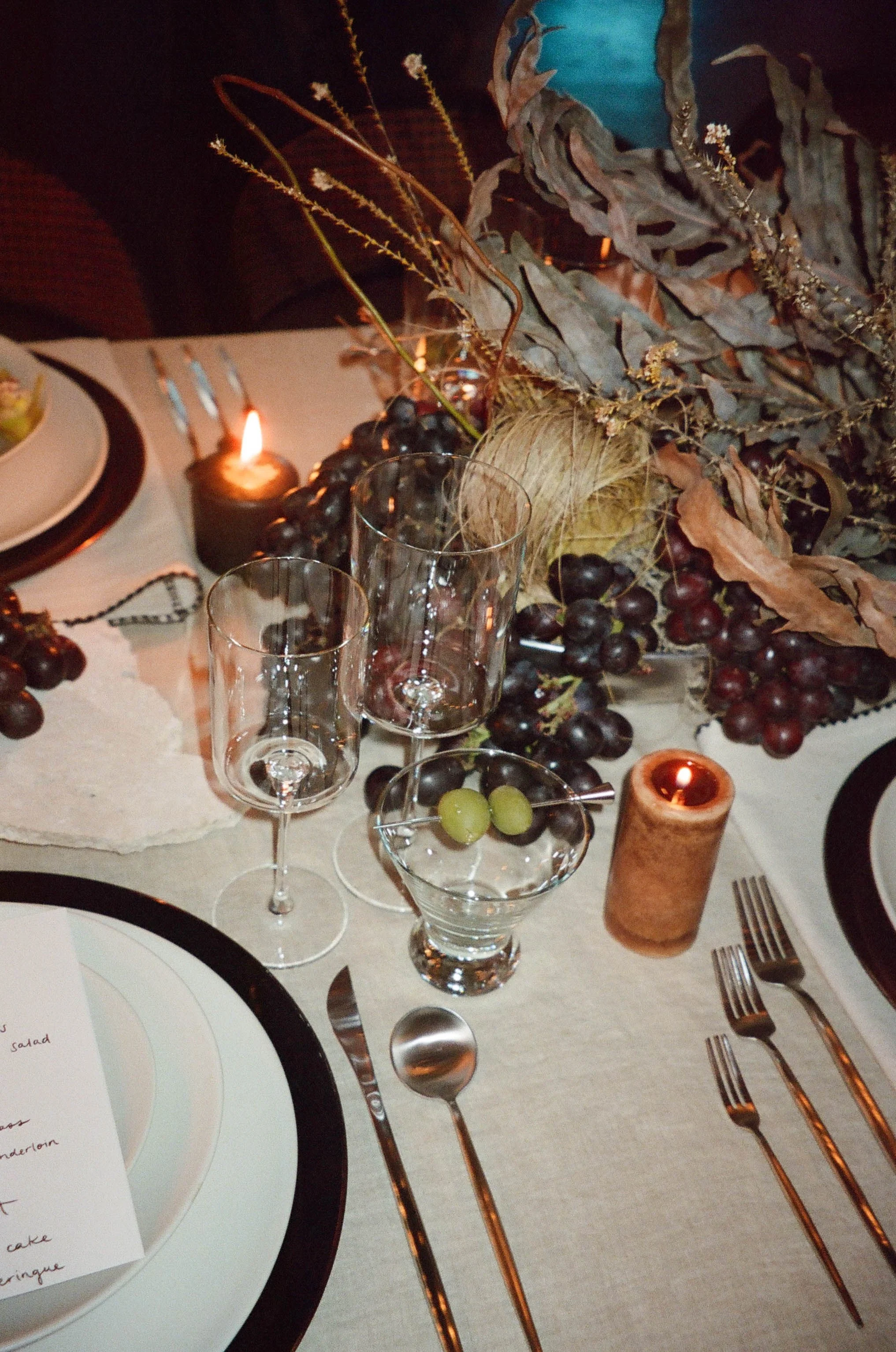 Elegant dining table with wine glasses, grape centerpiece, candles, and a menu card.
