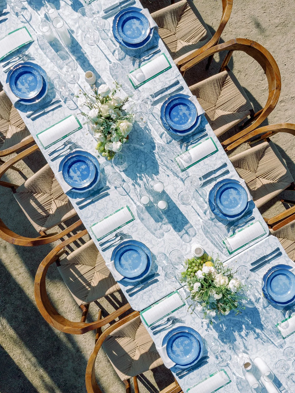 A long outdoor dining table set with blue and white dinnerware, glassware, silverware, floral centerpieces, and printed menus, shadowed by the sunlight.
