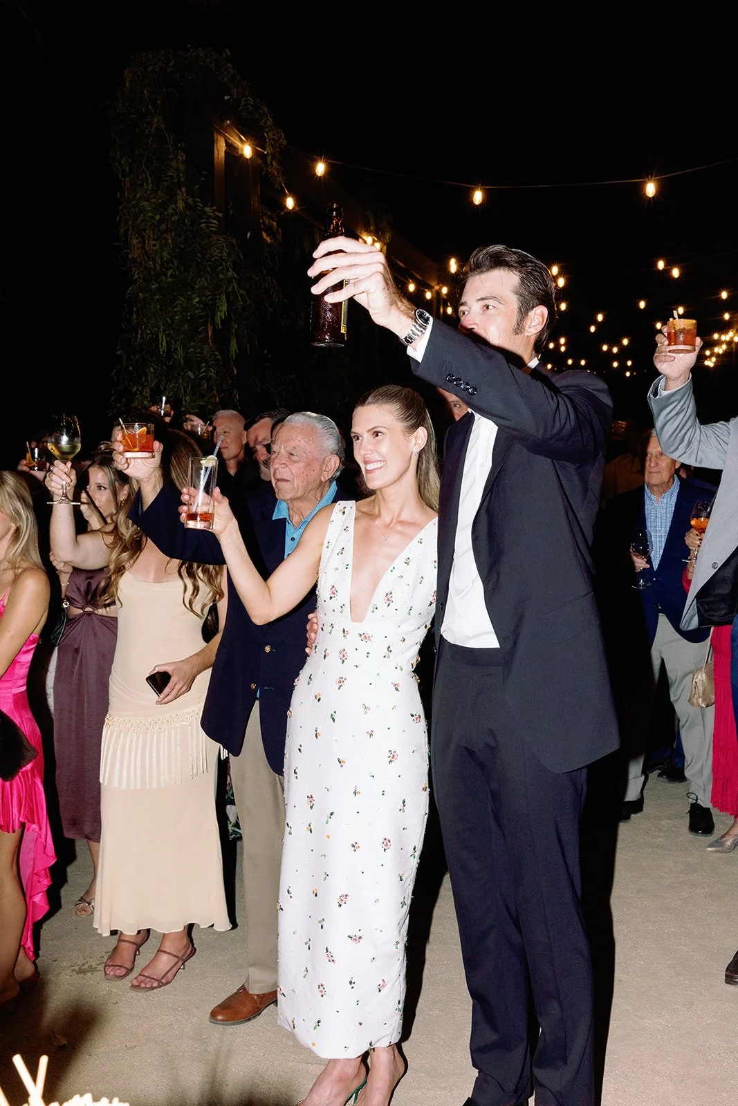 People celebrating at an outdoor event at night, raising drinks, with string lights overhead, including a smiling woman in a white dress and a man in a black tuxedo.