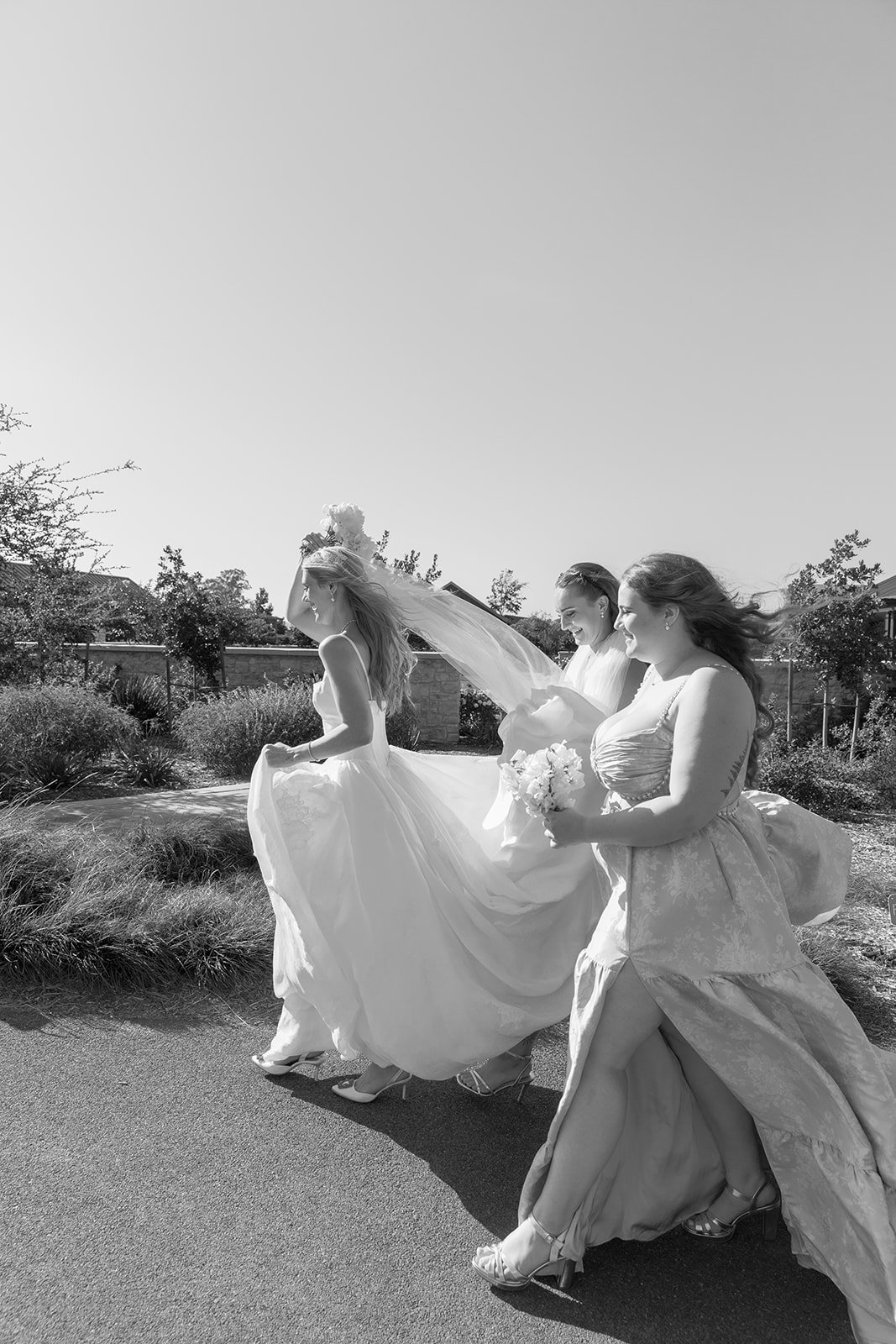 Three women in wedding dresses and veils walking outdoors on a sunny day, carrying bouquets of flowers, with wind blowing their hair and dresses.