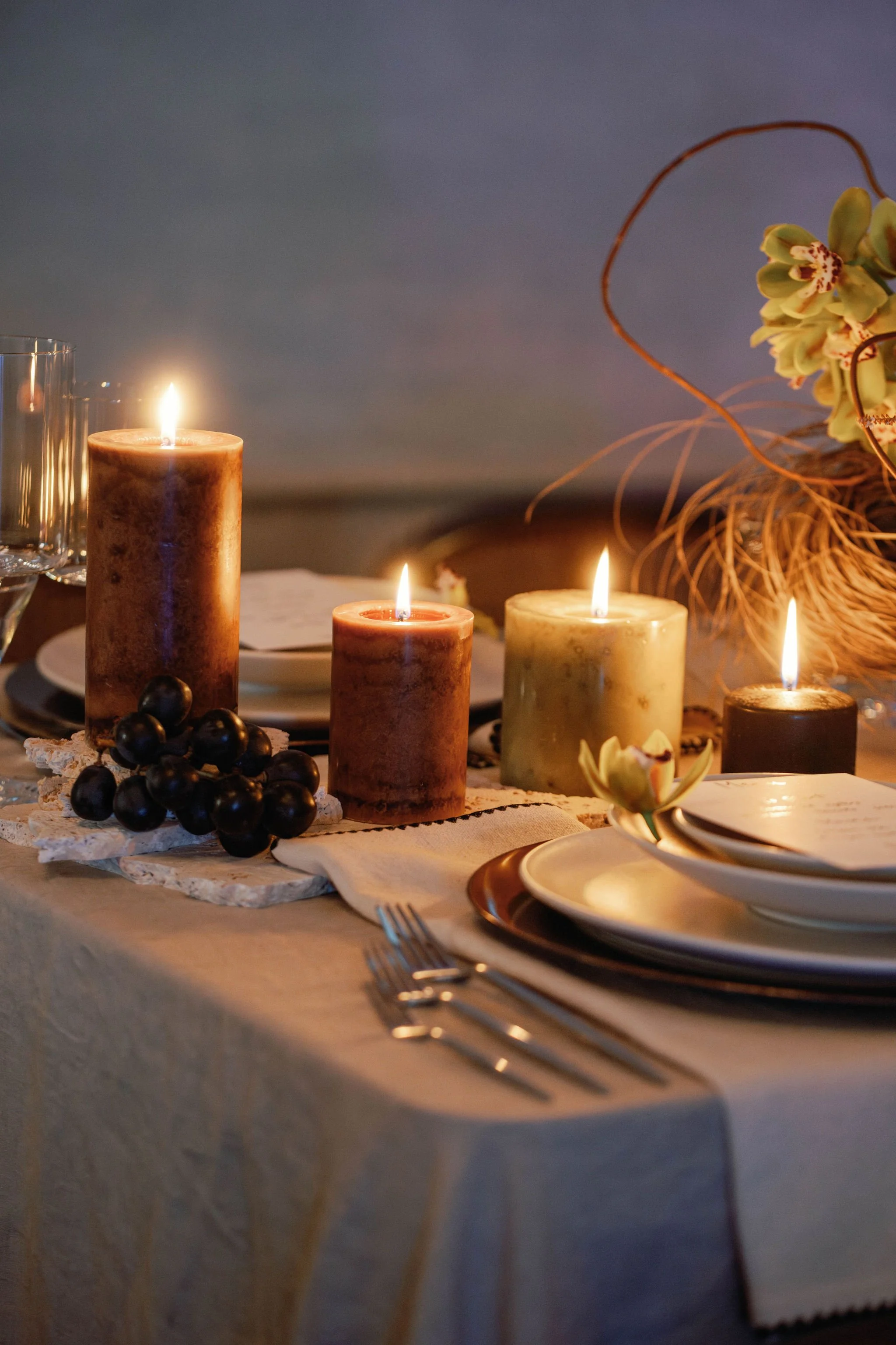 A dinner table decorated with four lit candles, grape cluster, plates, and cutlery, with a floral arrangement in the background.