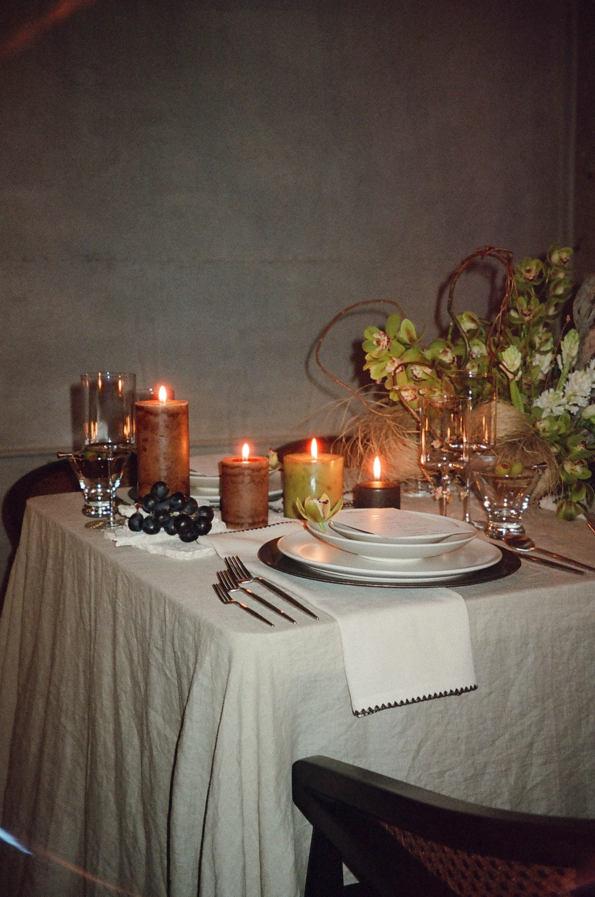 Decorative table setting with lit candles, wine glasses, plates, silverware, grapes, and floral arrangement.