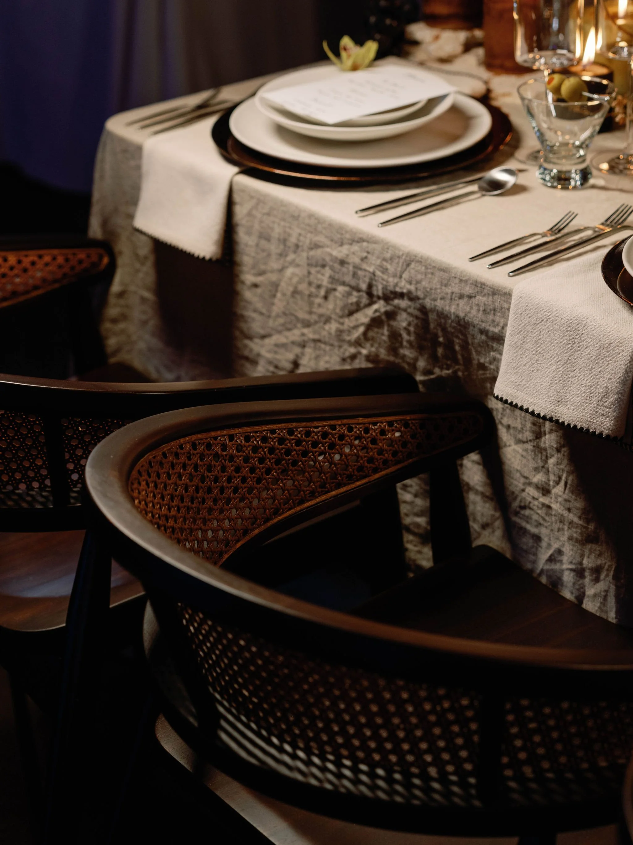 Elegant dining table set with white plates, silverware, wine glasses, and decorative flowers, surrounded by black and brown woven chairs.