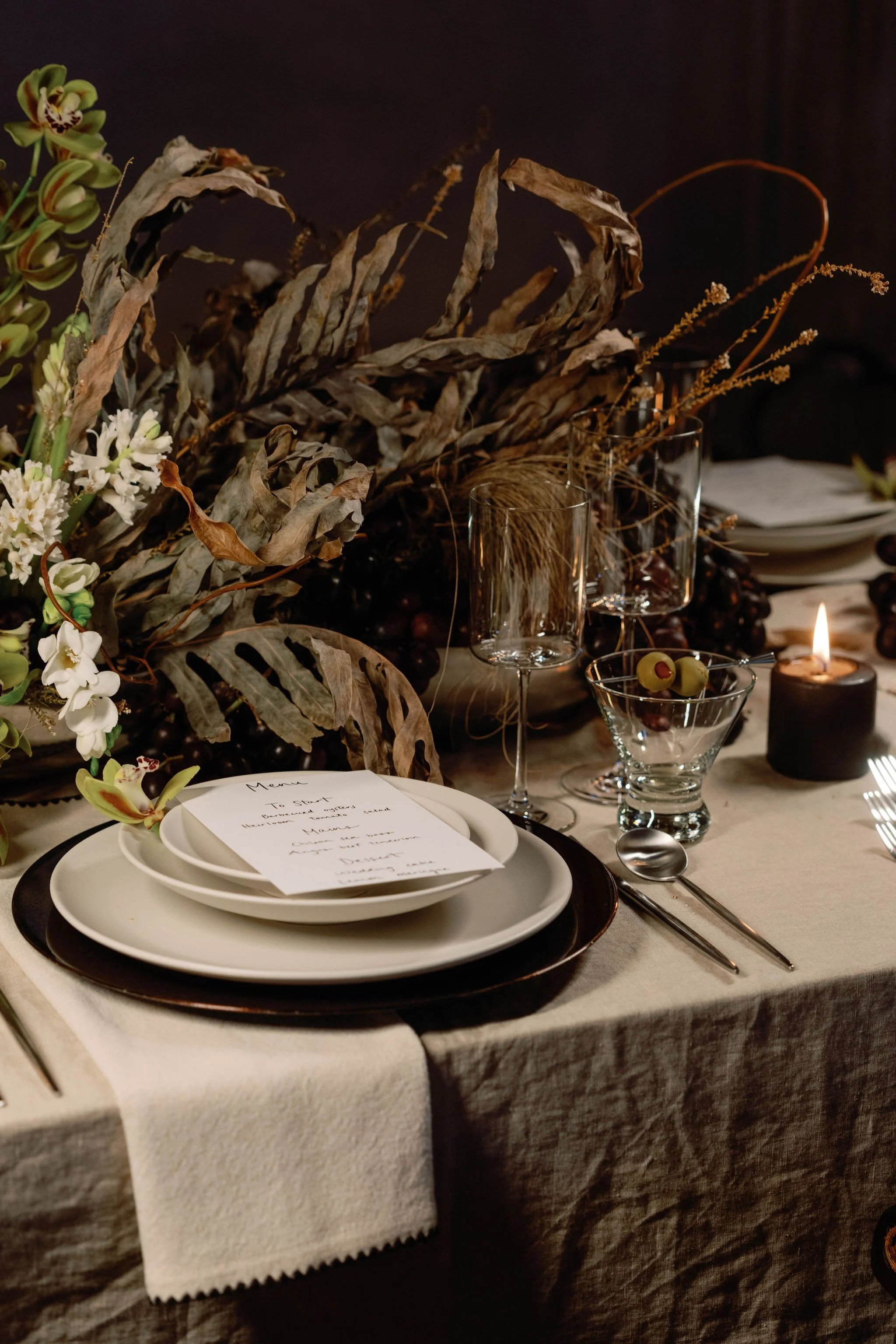 Table set for a formal dinner with white and black plates, wine glasses, a handwritten menu, silverware, a lit candle, and floral and dried leaf decorations.
