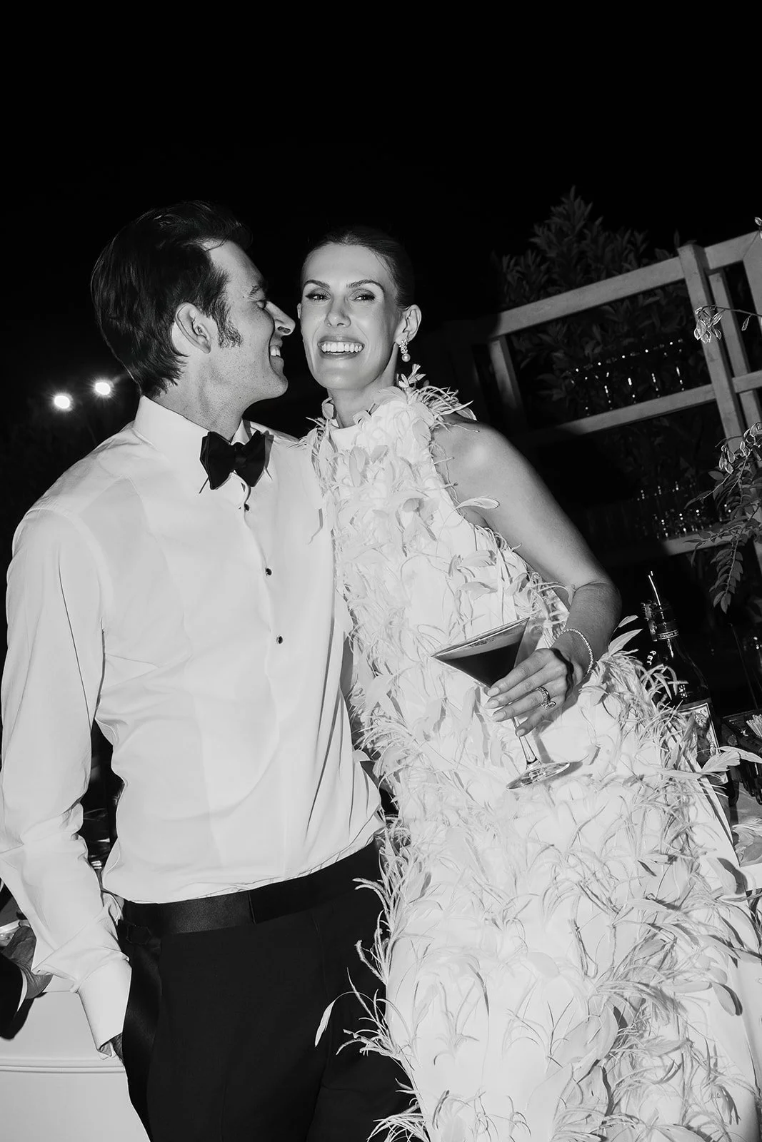 A black and white photo of a newlywed couple at their wedding reception, with the bride holding a cocktail glass and both smiling happily.