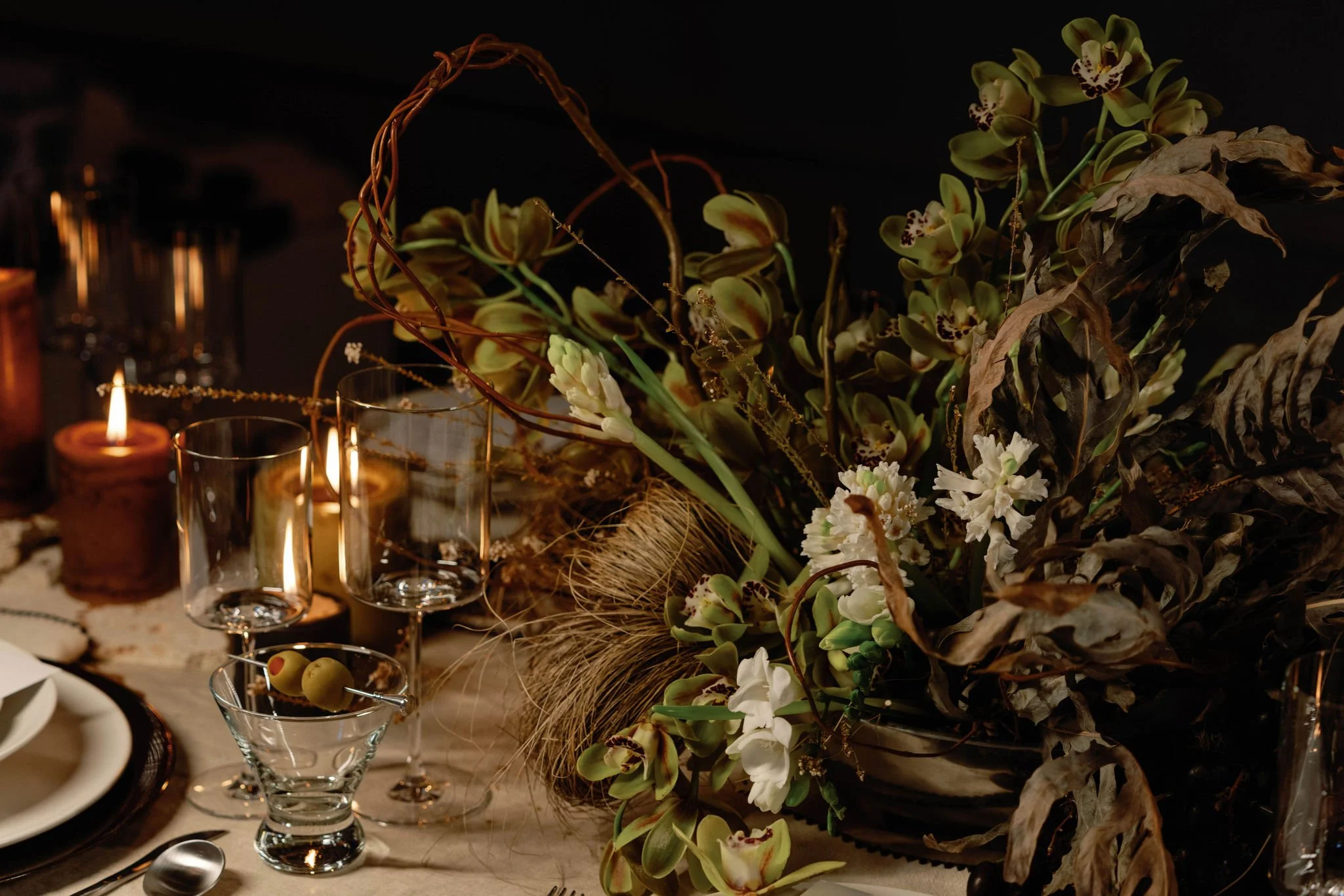 Decorative floral arrangement with orchids and dried leaves on a dinner table, lit by candles.