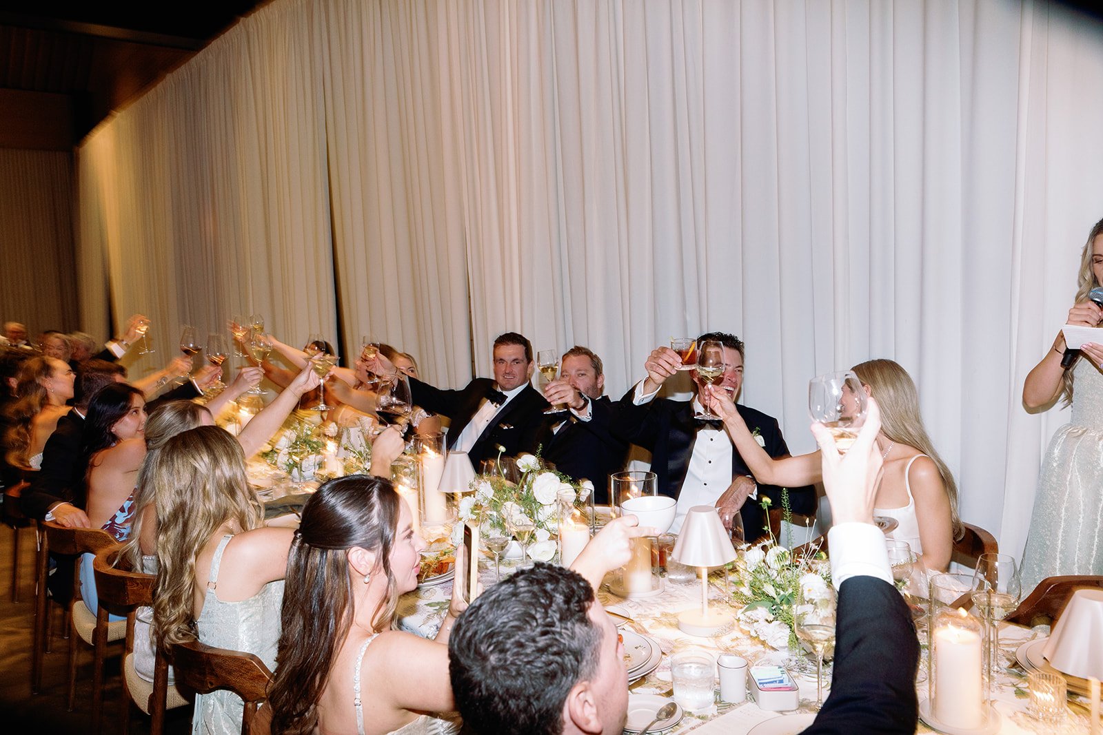 People at a wedding reception raising glasses for a toast, seated at a decorated table with white flowers and candles.