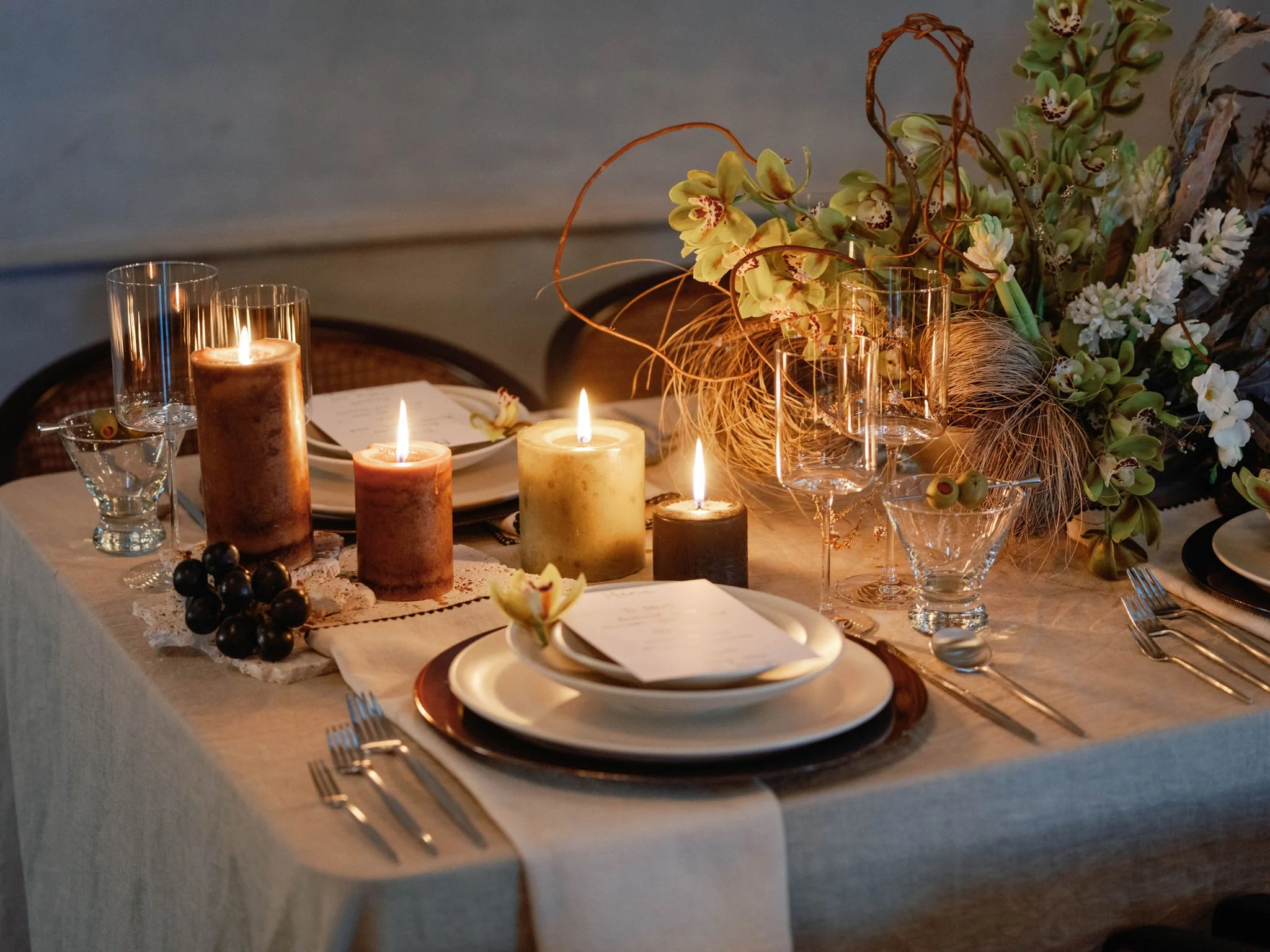 A table decorated with lit candles, empty wine glasses, plates with napkins and menus, a bunch of grapes, and a large floral centerpiece with green and white flowers and dried elements, set for a formal dinner.