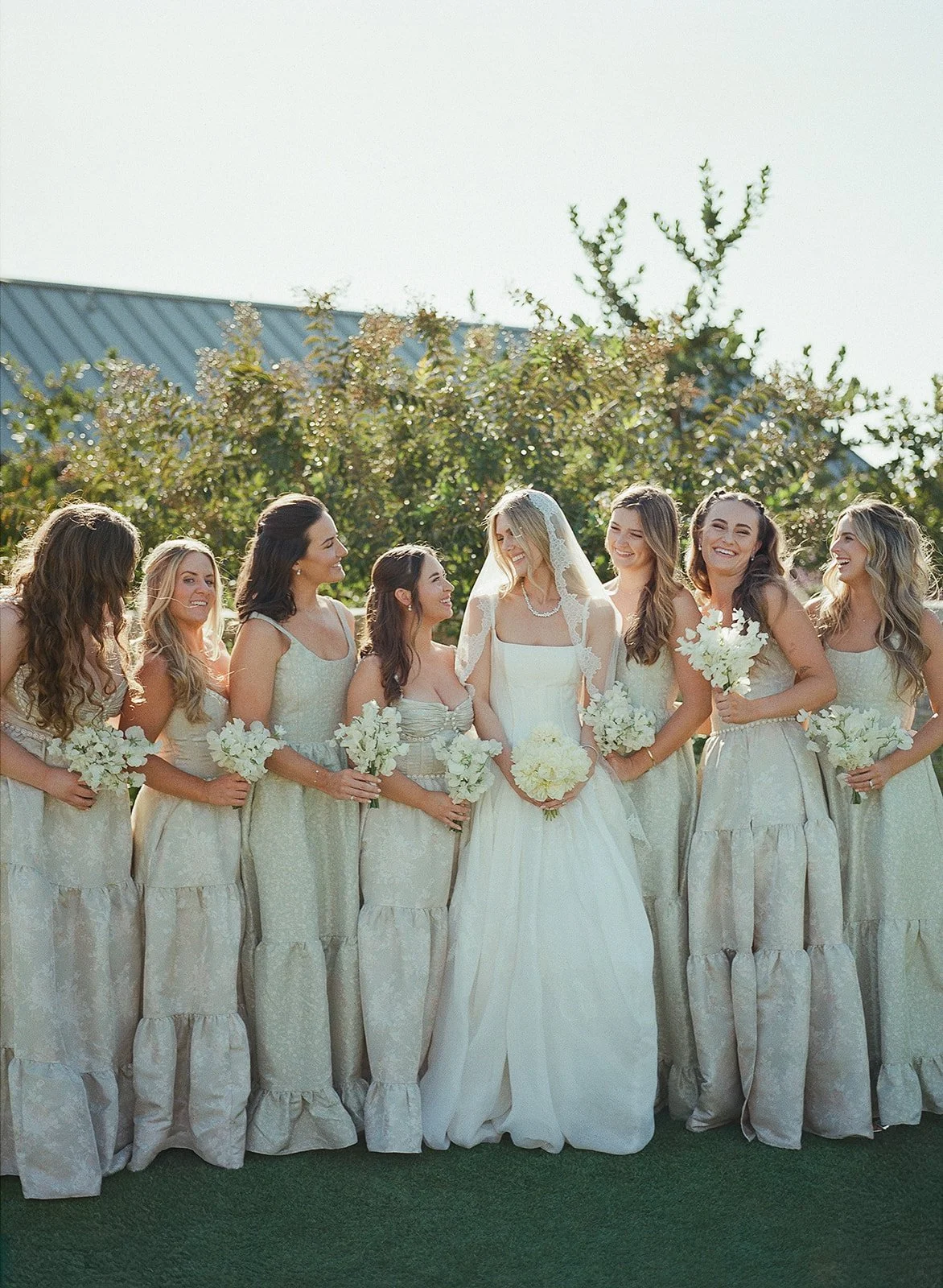 Group of seven women, including a bride in white wedding dress and veil, standing outdoors with greenery and a blue roof in the background, holding white floral bouquets.
