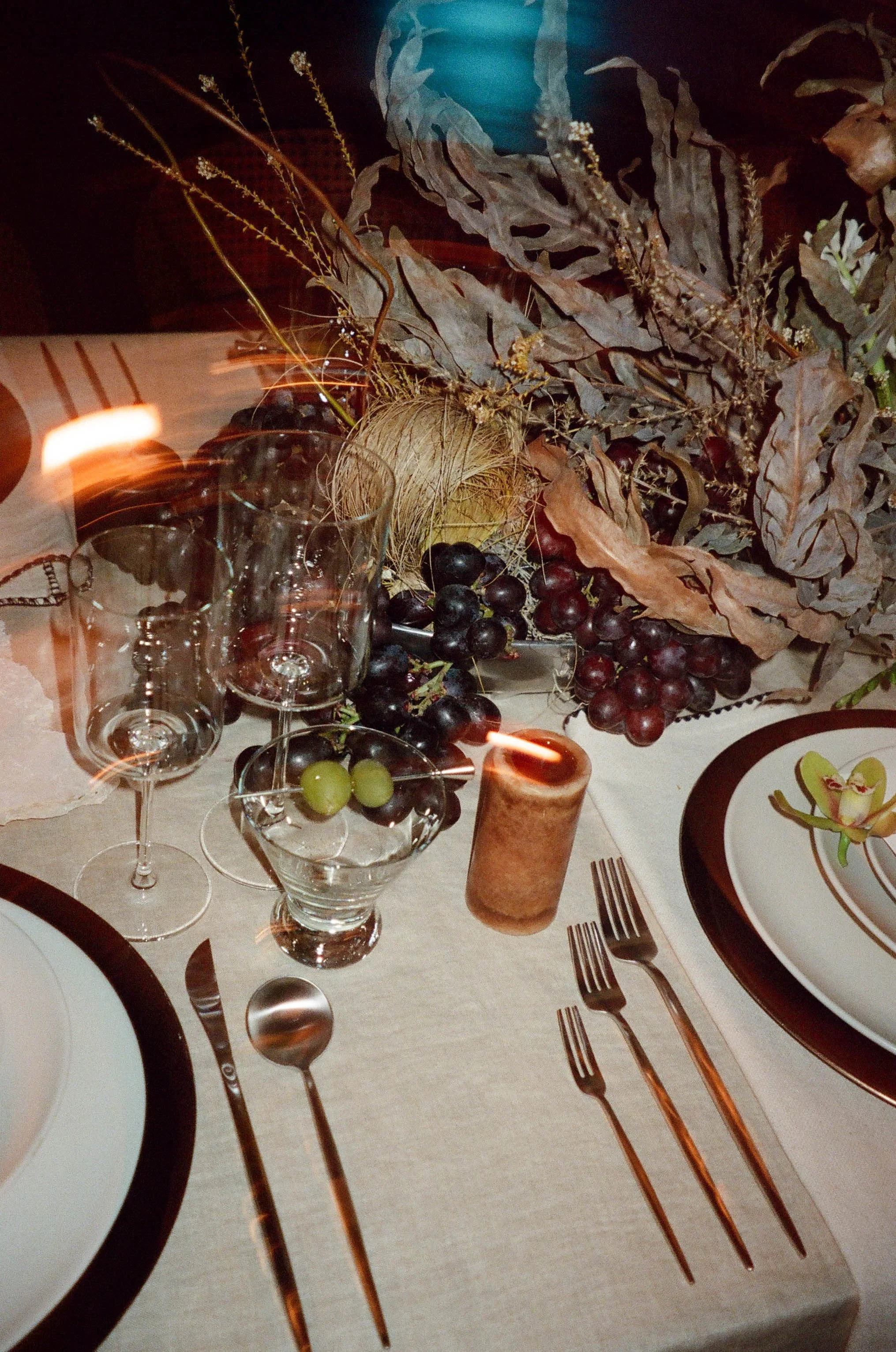 A formal table setting with plants, grapes, a candle, plates, forks, knives, and glasses on a table with a beige tablecloth.