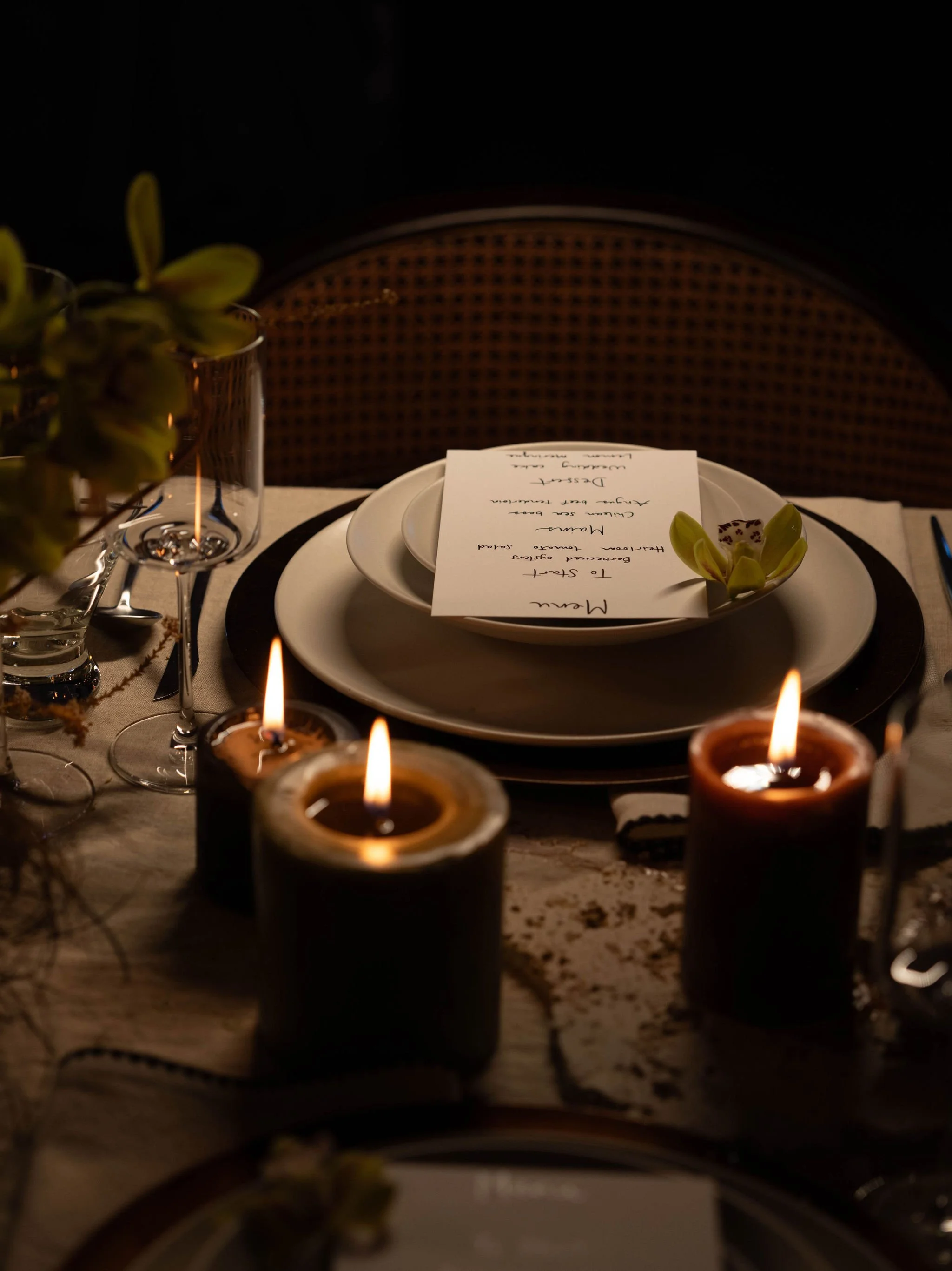 A dimly lit dining table with three lit candles, a dinner plate, a menu, and a wine glass, set for a romantic dinner with floral decorations.