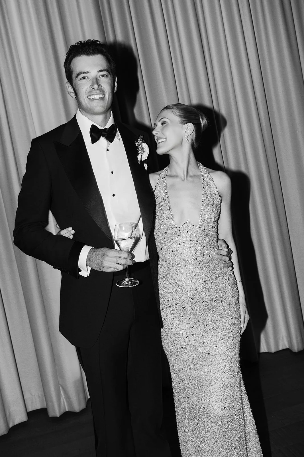 A man and woman in formal attire at an event, with the man holding a glass of wine, and both smiling at each other, standing in front of curtains.