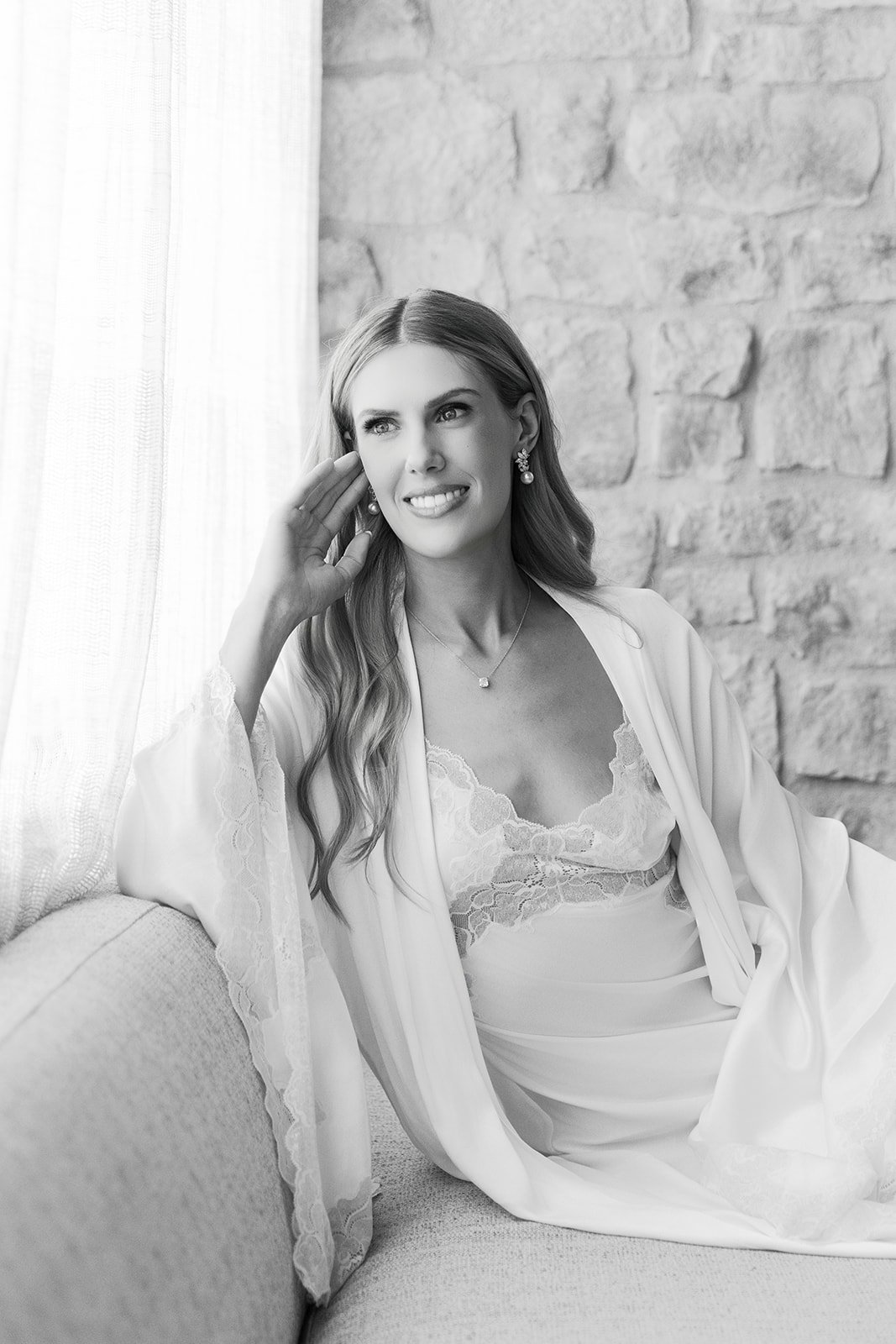 Black and white photo of a woman sitting on a sofa near a window, wearing a lace-trimmed satin robe, earrings, and a necklace. She has long wavy hair and is touching her face with her left hand, smiling gently.