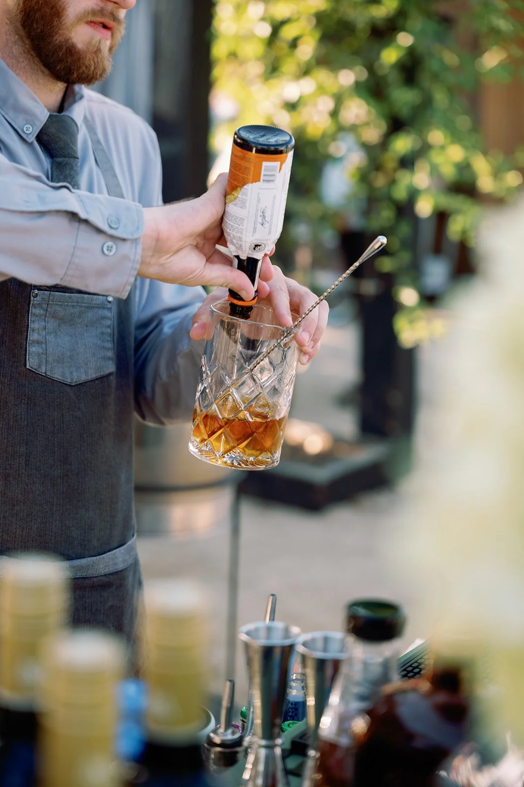 A bartender pours whiskey from a bottle into a glass with ice. The bartender wears a light blue shirt with rolled-up sleeves and a dark gray apron. The background is blurred with green foliage, and various bar tools and bottles are visible on the cou
