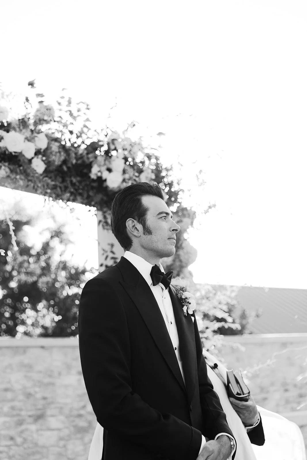 A black and white photo of a groom dressed in a tuxedo with a bow tie and boutonniere, standing outdoors with trees and a floral arch in the background.