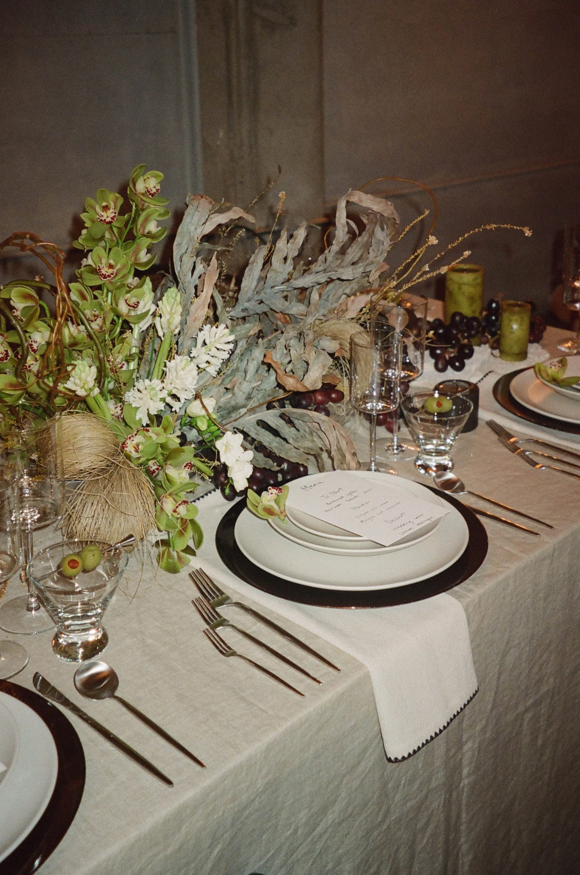 Elegant table setting with a floral centerpiece, white plates, silverware, wine glasses, a handwritten menu, and green candles on a beige tablecloth.