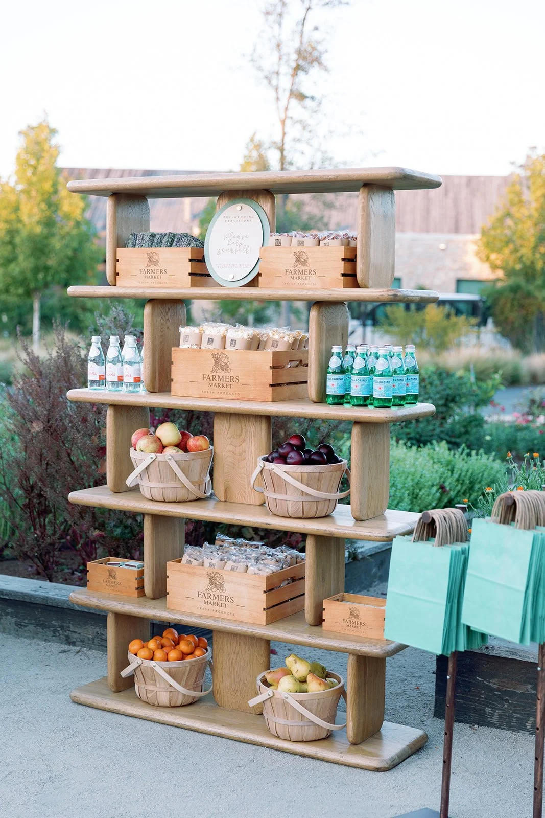 Outdoor display stand with wooden shelves holding apples, bottled water, and small boxes of goods, set in a garden or park.