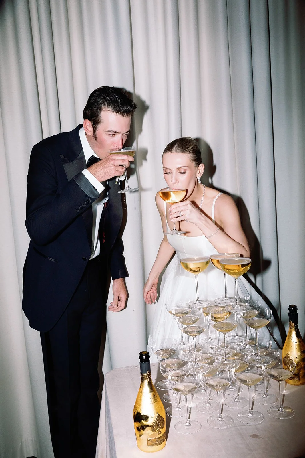 A bride and groom in wedding attire are drinking champagne from large glasses. The bride is wearing a white dress and jewelry, while the groom is in a dark tuxedo. There is a tower of champagne glasses and two bottles of champagne on the table.