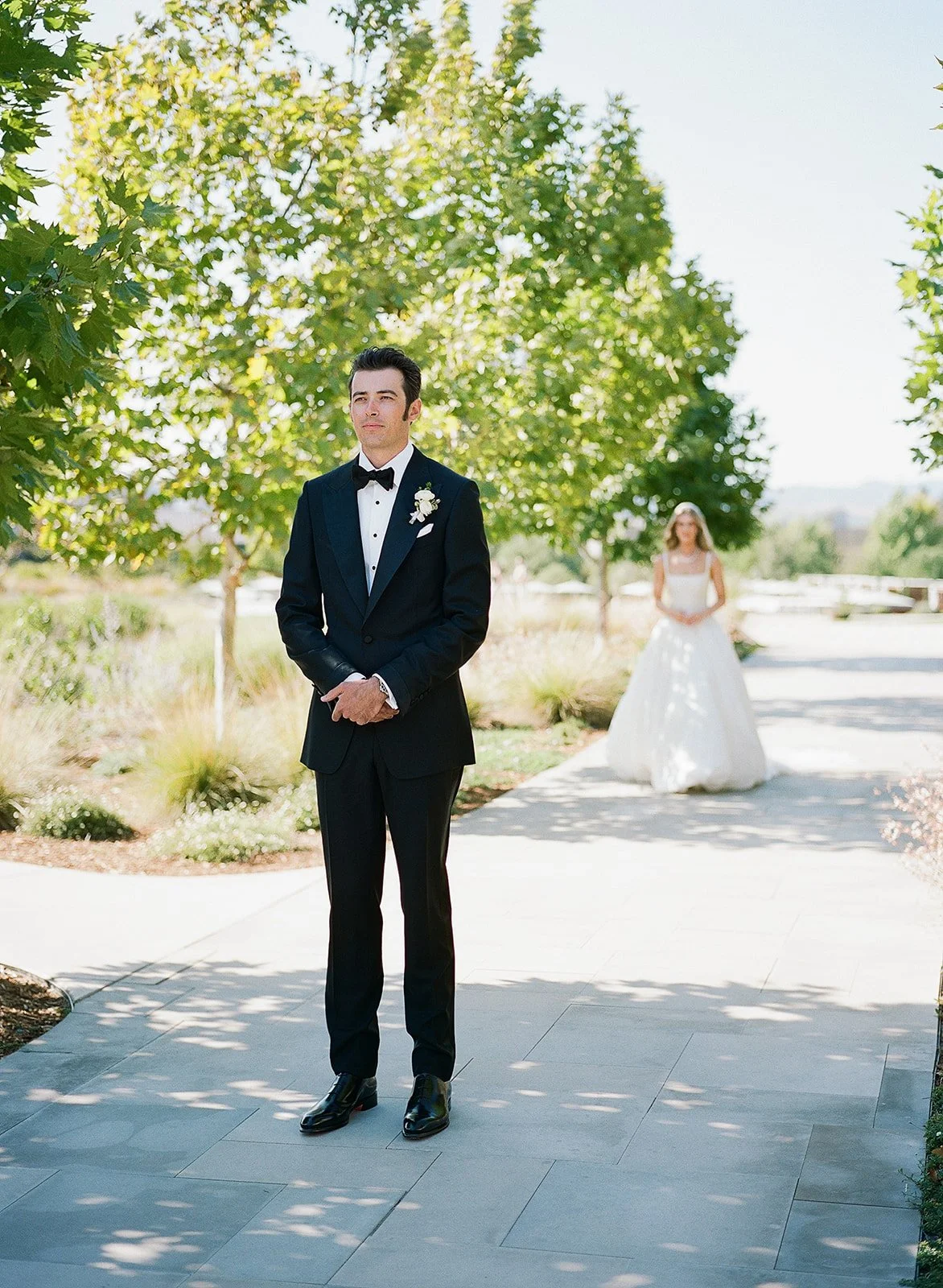 A groom in a black tuxedo with a white shirt and black bow tie stands outdoors on a paved walkway, looking thoughtful, with a bride in a white wedding dress in the background, standing under a tree with green foliage on a sunny day.