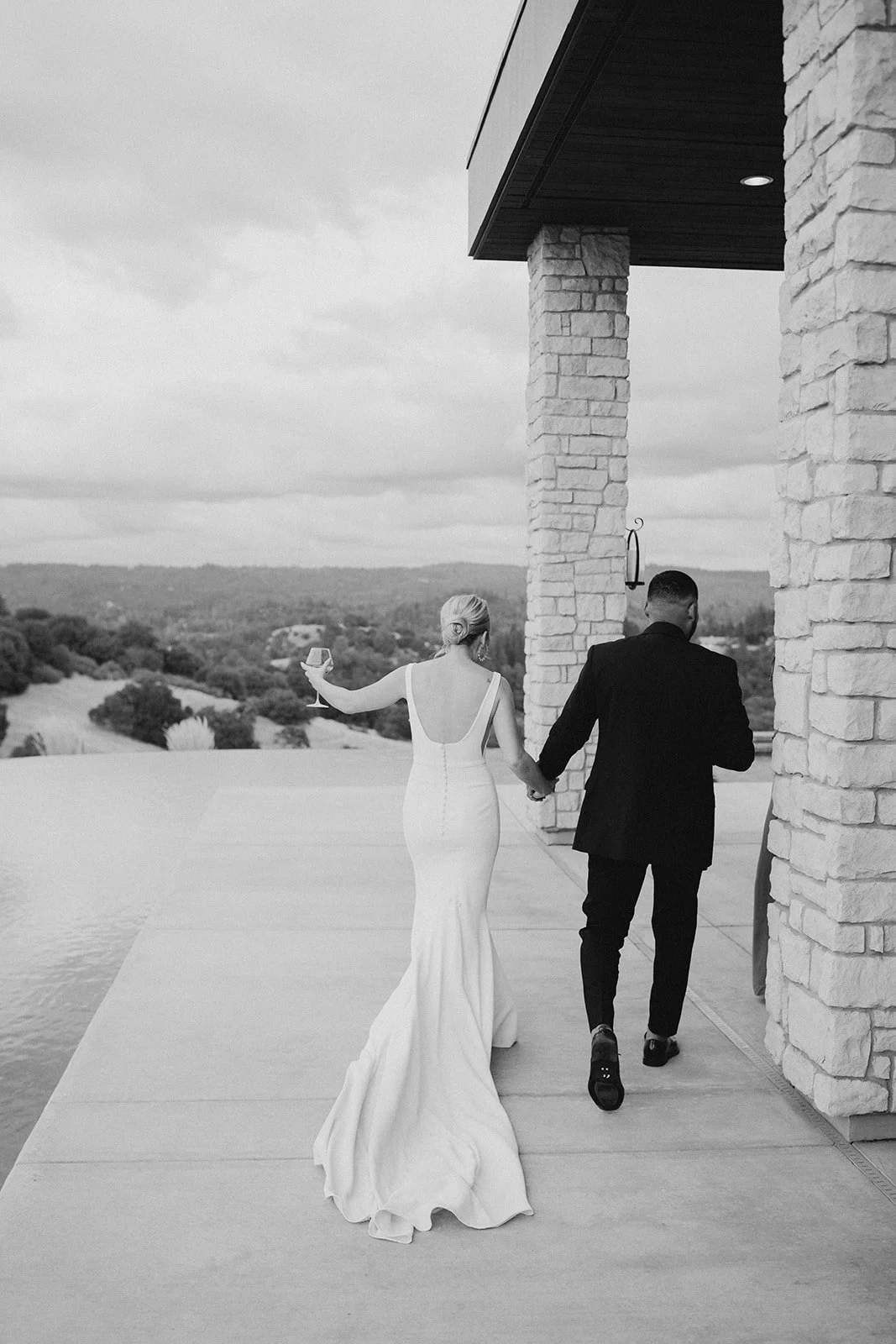A black and white photo of a bride and groom walking hand in hand on a patio, with the bride holding a glass of wine, overlooking a scenic landscape.