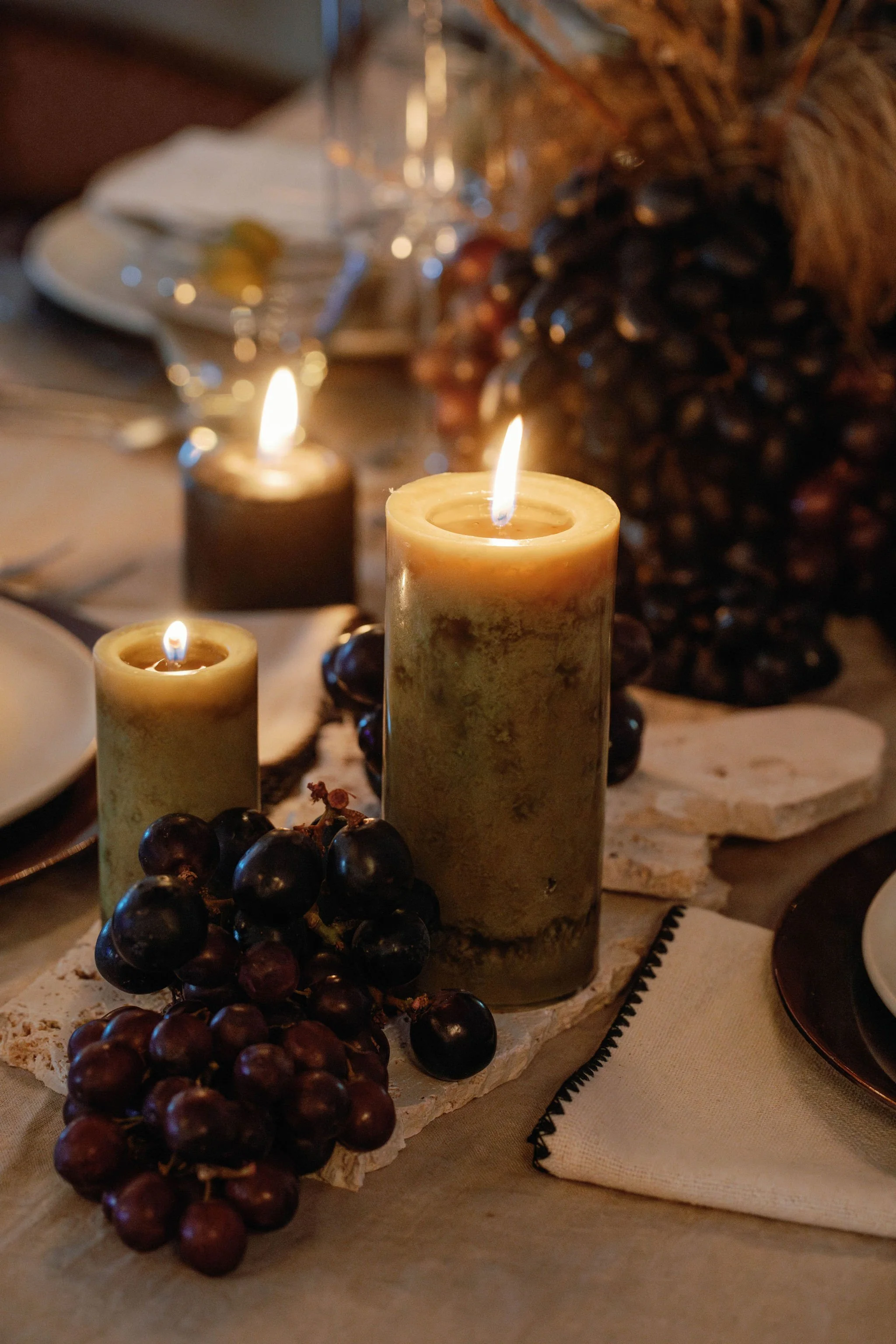 A candlelit table setting with two candles, clusters of dark grapes, stone slabs, and a blurred background of a table with plates and glassware.