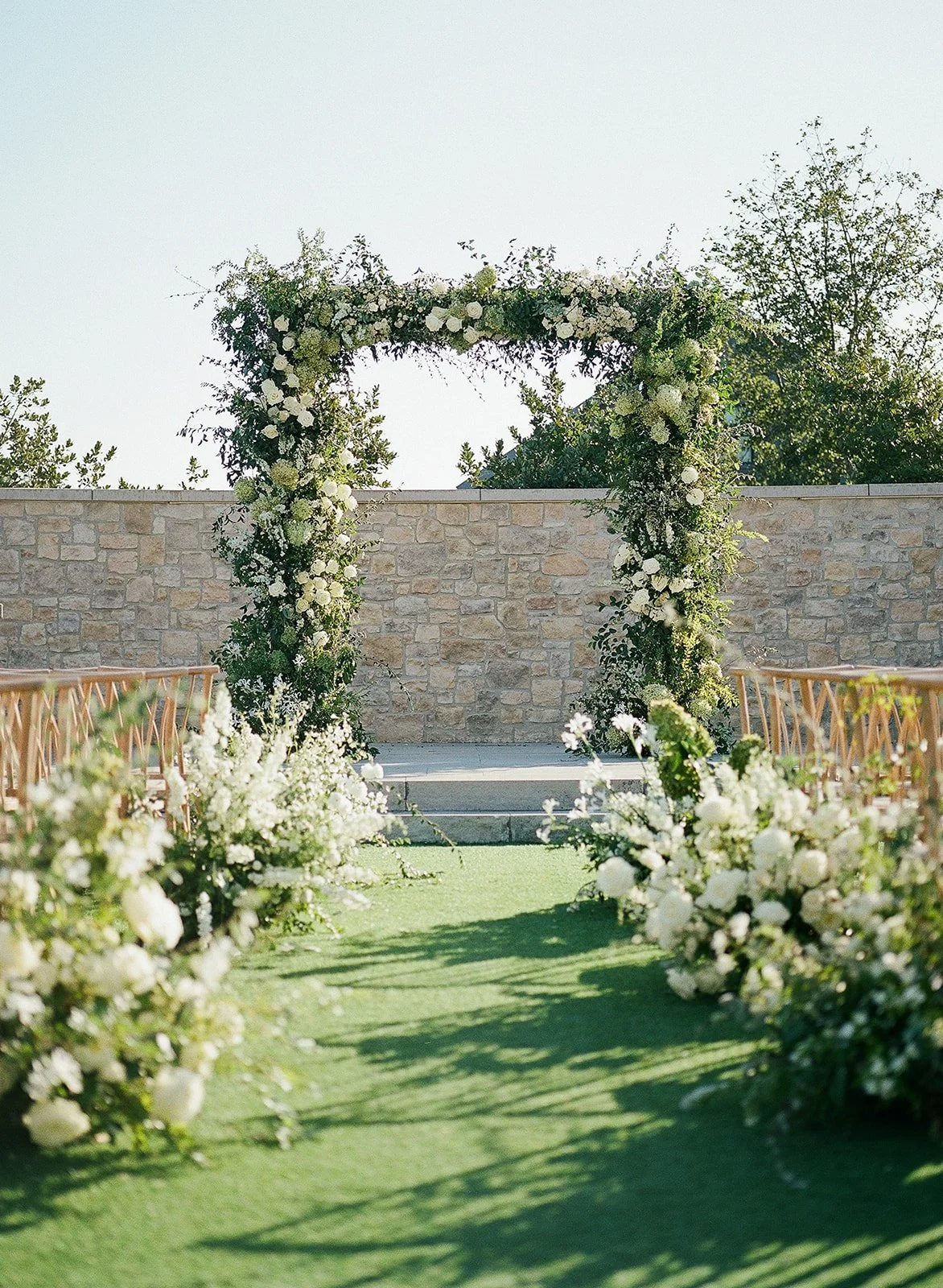 Outdoor wedding ceremony setup with a floral arch and white floral arrangements along the aisle on a green carpet, with chairs on both sides and a stone wall in the background.