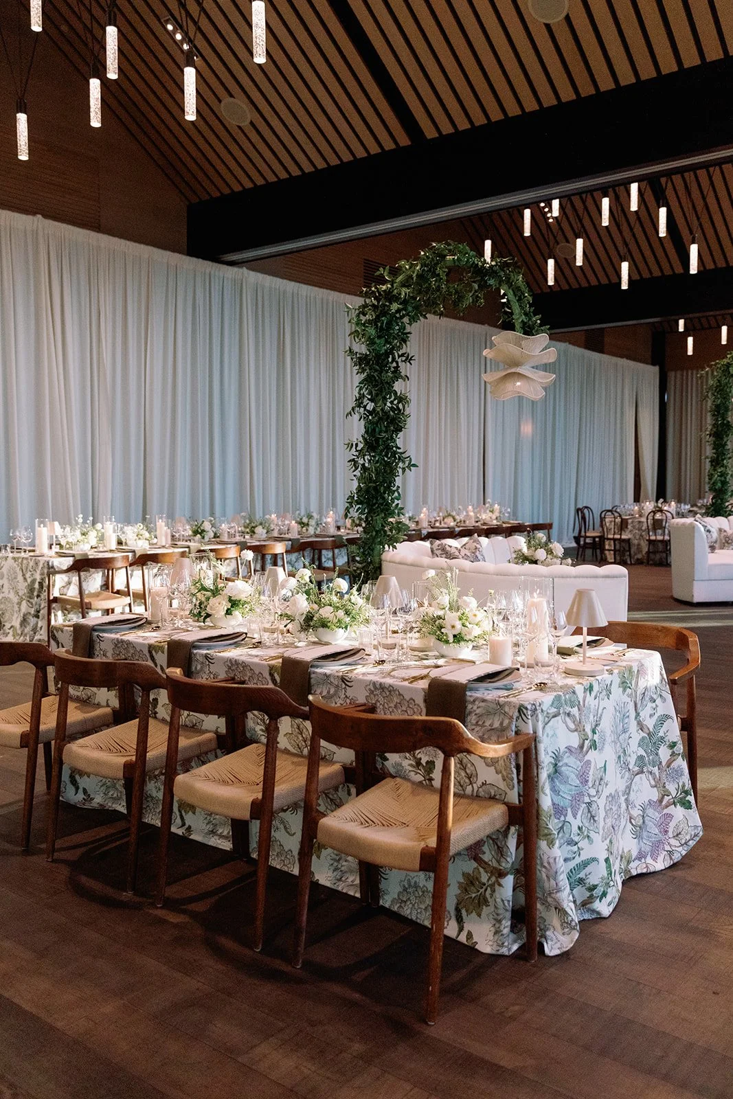 Elegant indoor event setup with a large round table decorated with white floral arrangements, candlelit tableware, and a floral and greenery arch overhead in a modern venue with warm lighting and wooden ceiling.
