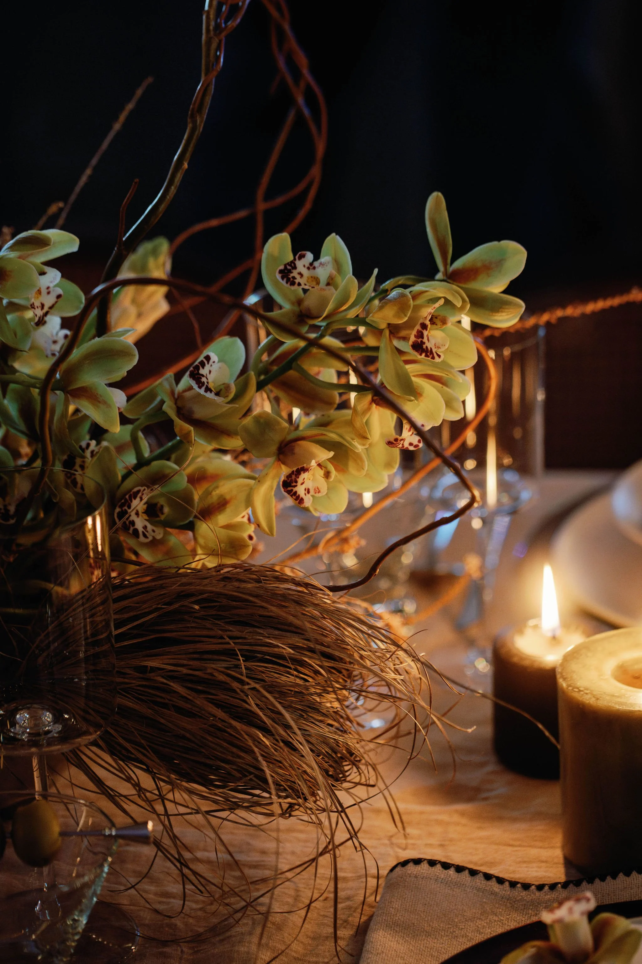 A table decorated with a bouquet of green orchids, a glowing candle, and elegant glassware in a dimly lit setting.