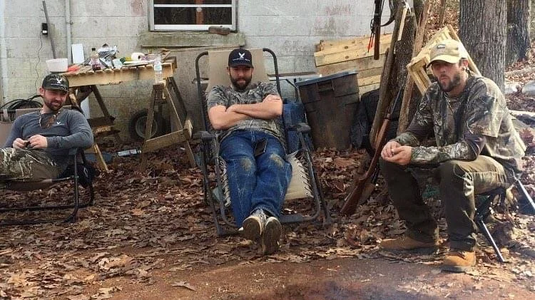Three men sitting outdoors with fallen leaves and wooden pallets, with the three dove brothers sitting in a chair, in front of a house with a window and a workbench in the background.