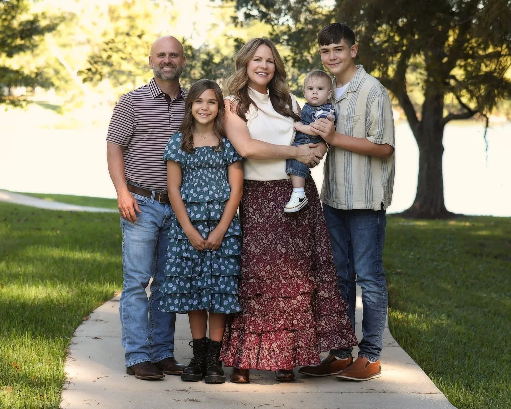 Family of six standing on a sidewalk near a lake with trees in the background, smiling at the camera.