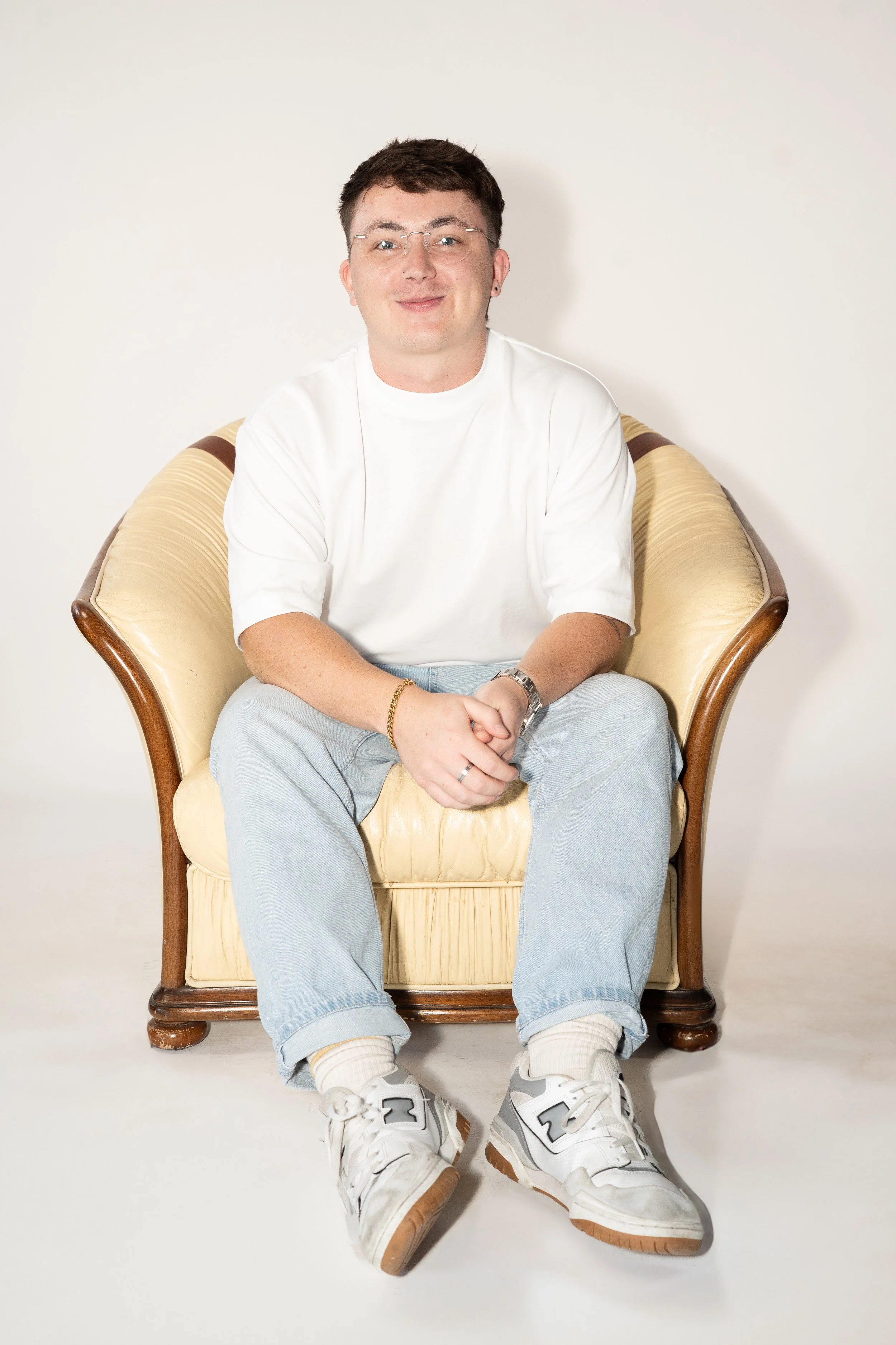 Young man sitting in a beige armchair against a plain light-colored wall, smiling, wearing glasses, a white t-shirt, light blue jeans, and white sneakers.